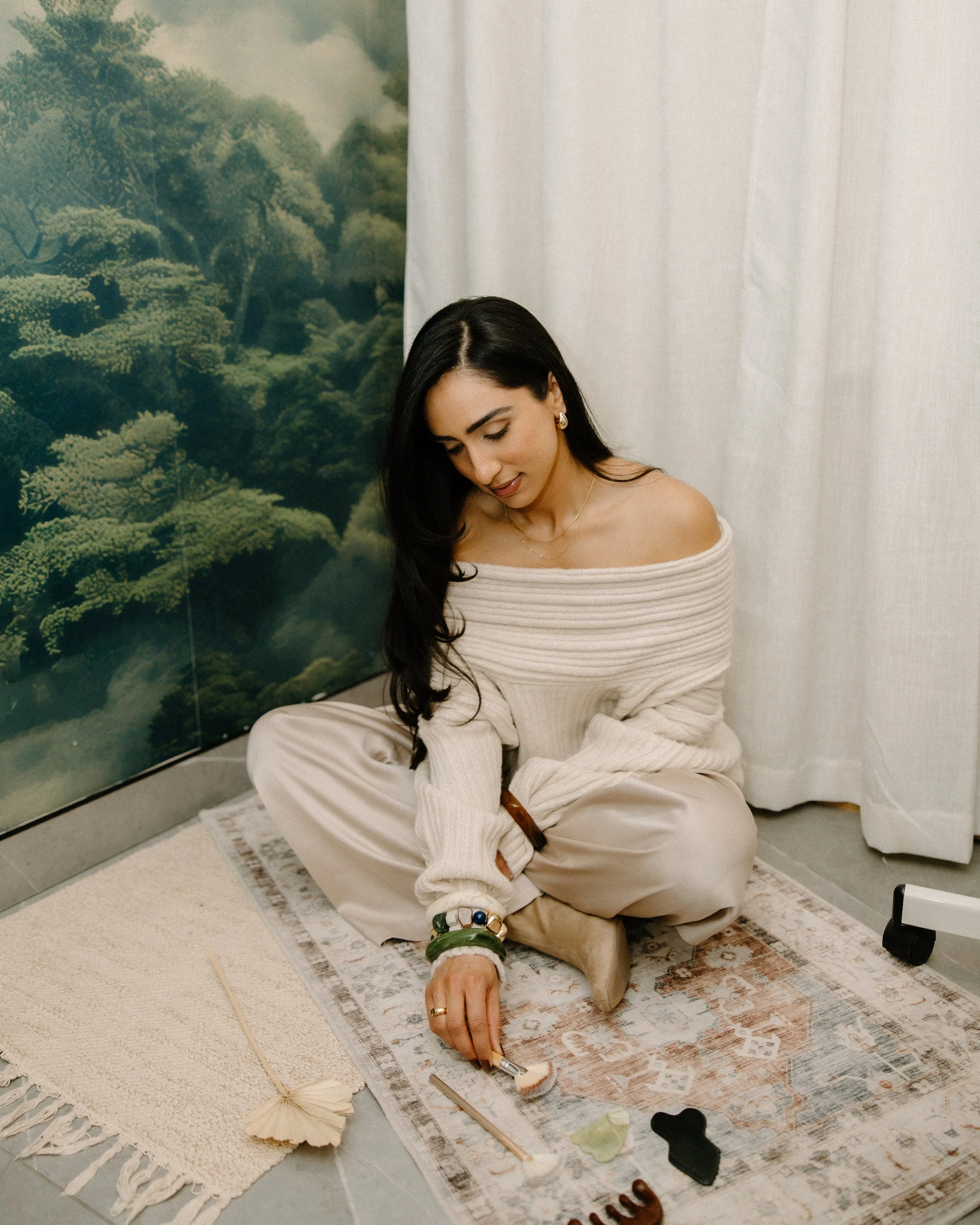 A woman with long dark hair, dressed in a beige off-shoulder top and beige pants, is sitting cross-legged on a patterned rug inside a room. She is painting or drawing on a large sheet of paper with various art supplies scattered around her, including brushes, colored stones, and decorative objects. There is a white curtain and a wall with a large painting of a lush green landscape behind her.