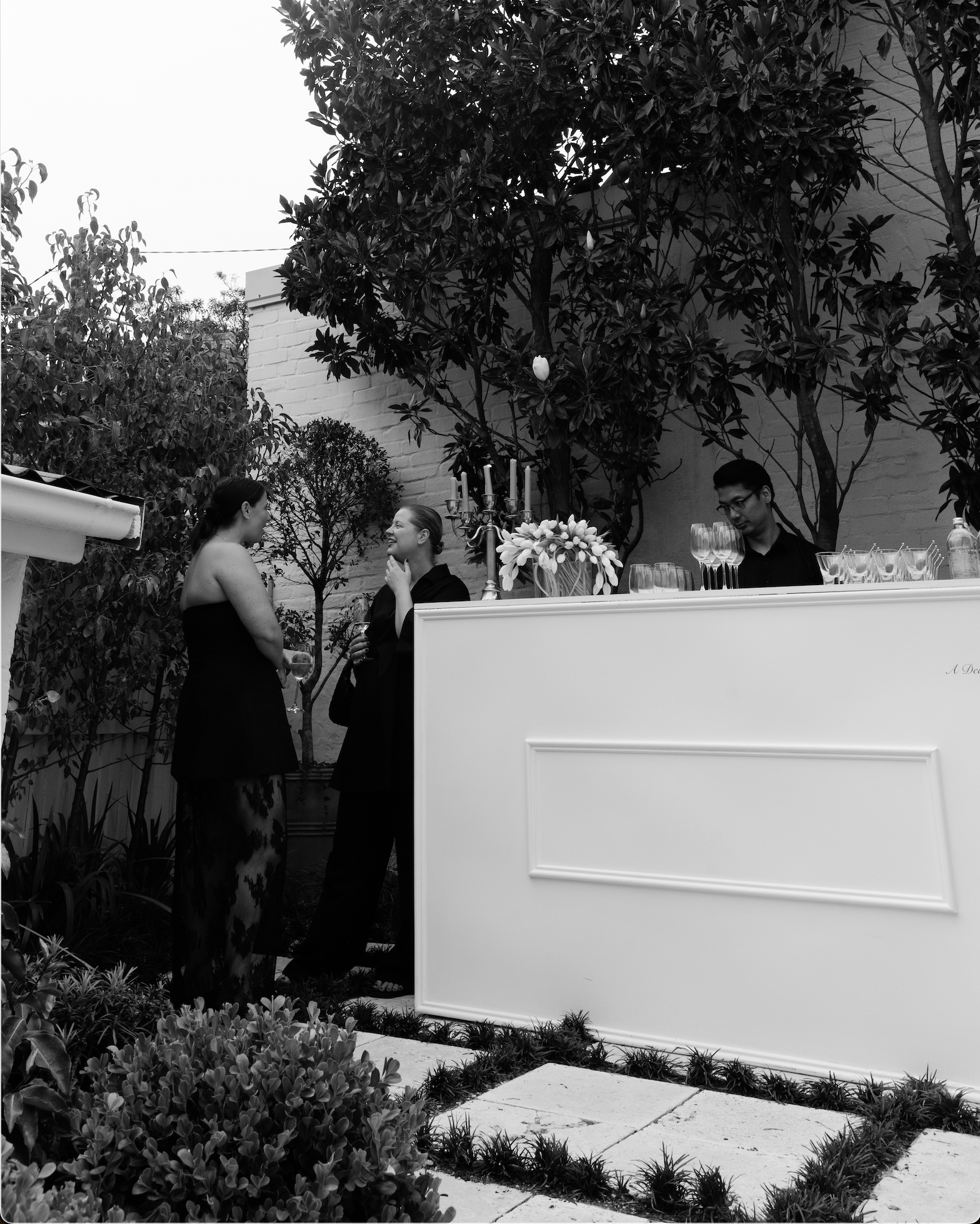 Two women in black dresses talking near an outdoor bar with a bartender, a man, and floral decorations, surrounded by trees and plants.