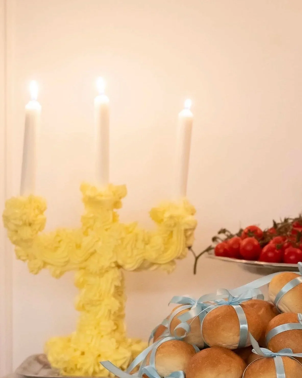 A yellow menorah made of candles with three lit candles on top, decorated with yellow flowers, a plate of tomatoes, and a pile of round bread rolls tied with blue ribbons on a table.
