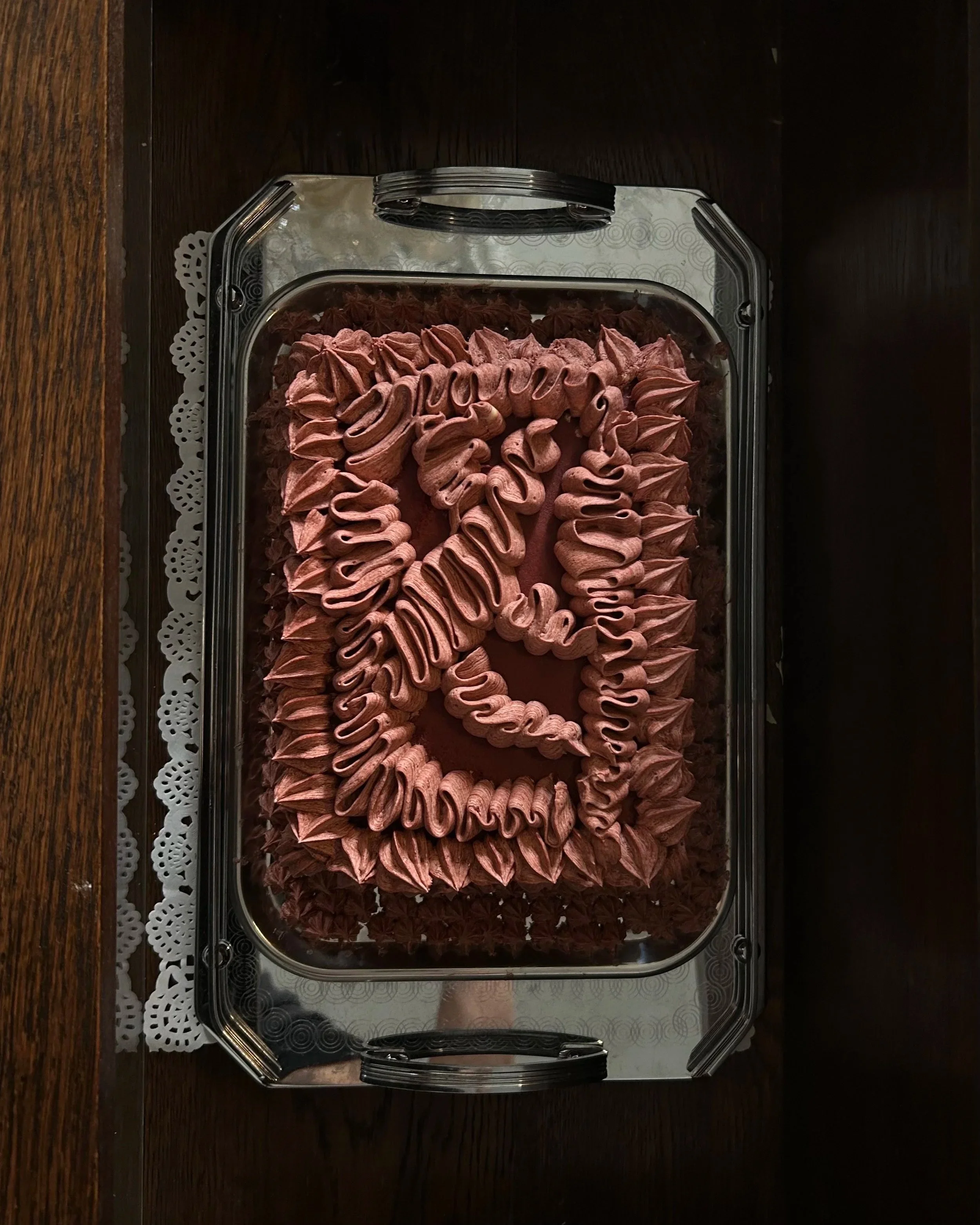 A rectangular chocolate cake with pink frosting writing on top that says 'Happy Birthday.' The cake is decorated with pink frosting piped around the edges, placed on a silver tray.