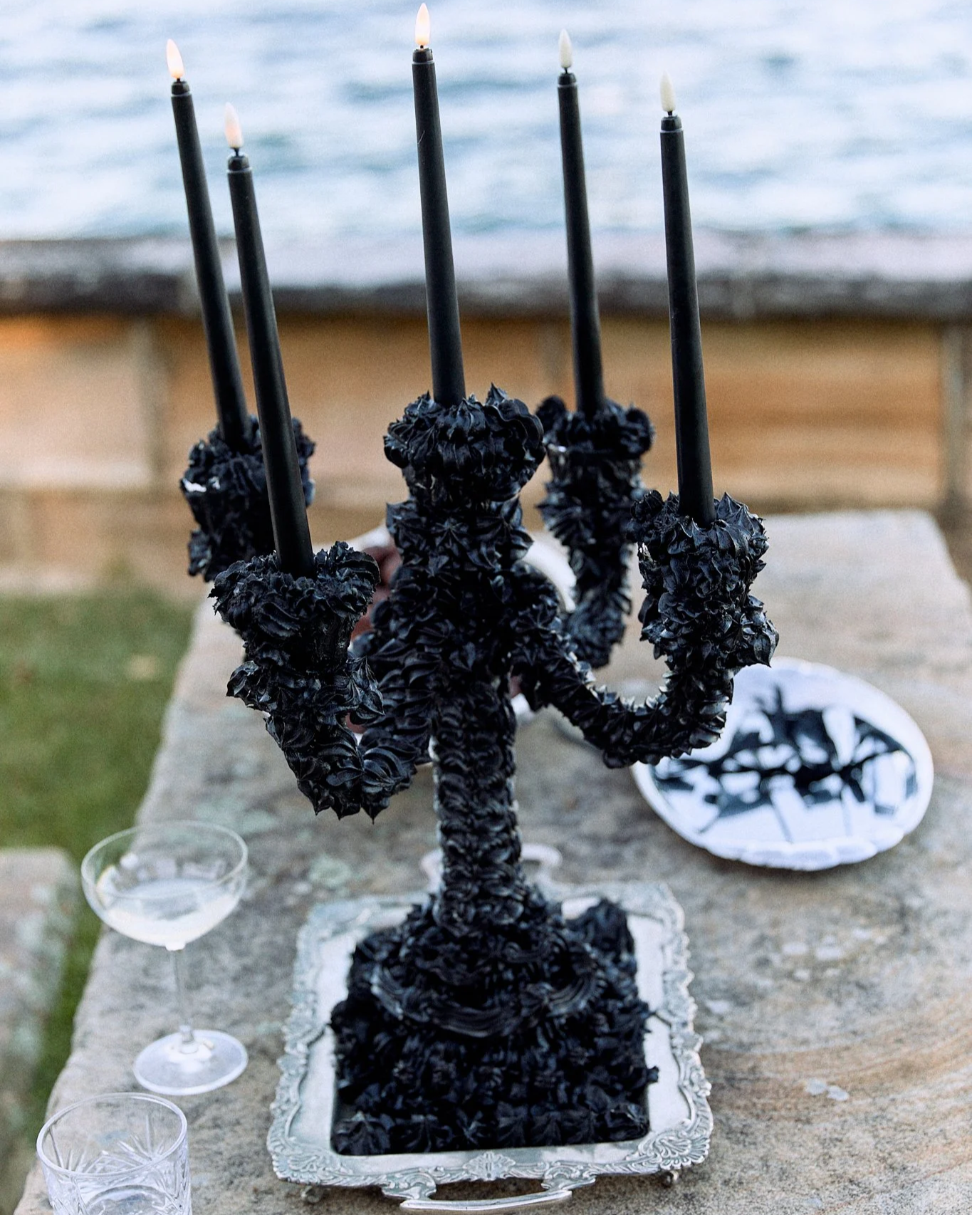 A black candelabrum with multiple lit candles, decorated with black ruffled material, placed on a silver tray on a wooden table outdoors near water.