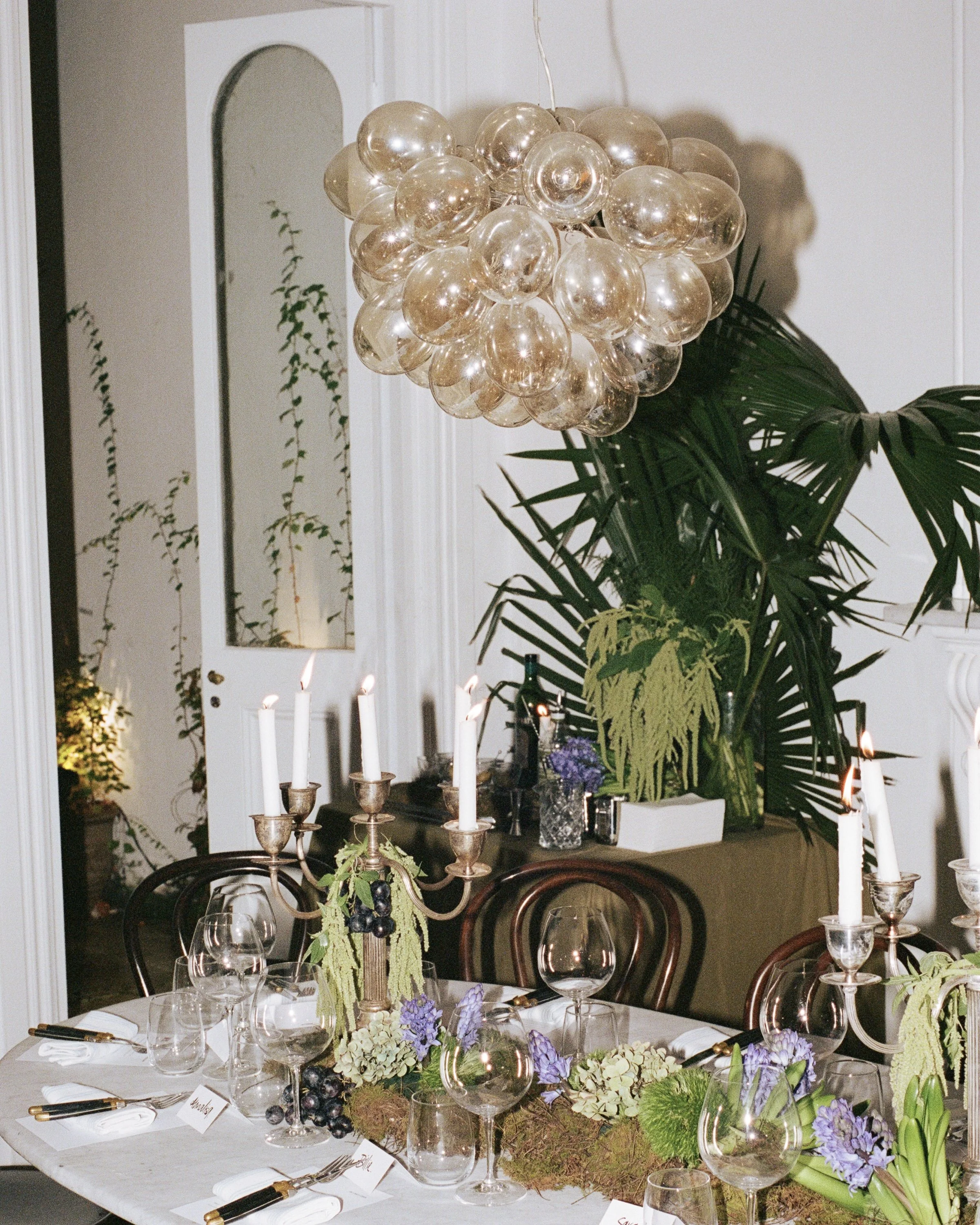 A decorated dining table with floral arrangements, glasses, plates, candles, and a modern chandelier made of glass bubbles hanging above, in a white-walled room with greenery.