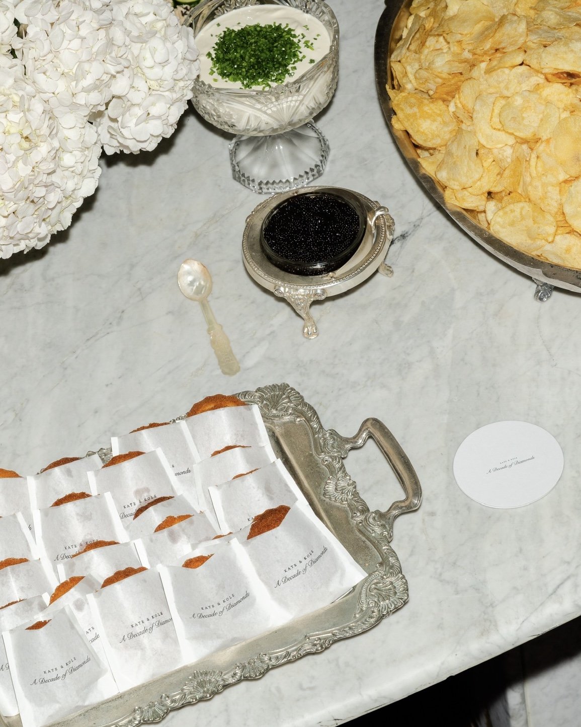 A marble table setting with white hydrangea flowers, a bowl of cream with chopped chives, a dish of black caviar, a silver tray with packaged mini muffins labeled 'A Decade of Diamonds,' a small spoon, a larger serving dish of potato chips, and a white oval card.