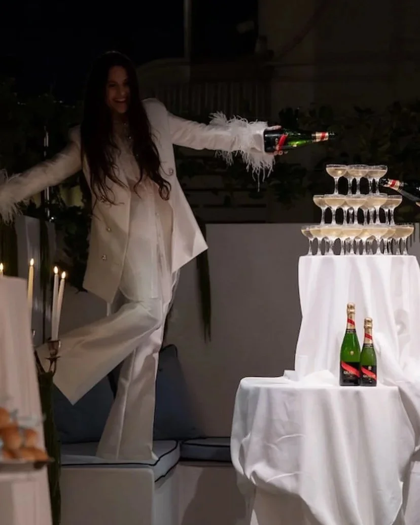 Woman in white suit pouring champagne into a pyramid of glasses at a celebration event.