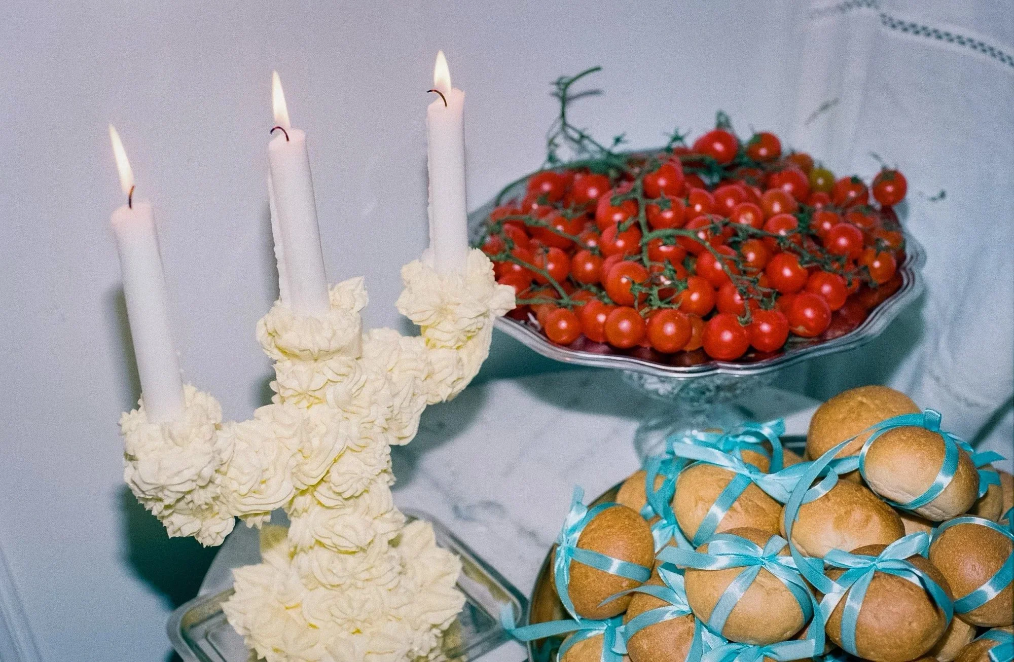 Decorative table with three lit candles on a floral arrangement, a bowl of cherry tomatoes on a stem, and a plate of small baked goods tied with blue ribbons.