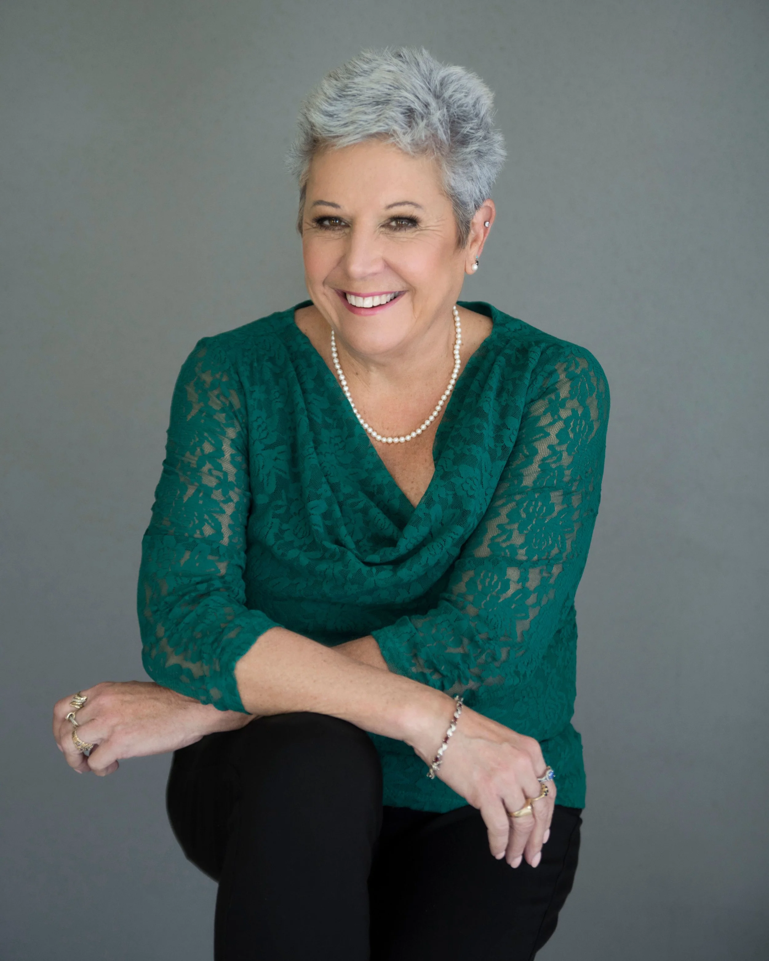 A smiling middle-aged woman with short gray hair, wearing a teal lace top, a pearl necklace, and earrings. She is sitting against a plain gray background, with her arms crossed and hands resting on her knee.