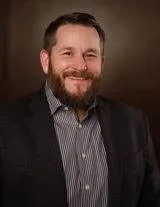 A woman with blonde hair wearing a black top, a tan blazer, and a turquoise necklace, smiling at the camera, seated at a table with a decorative wooden panel in the background.