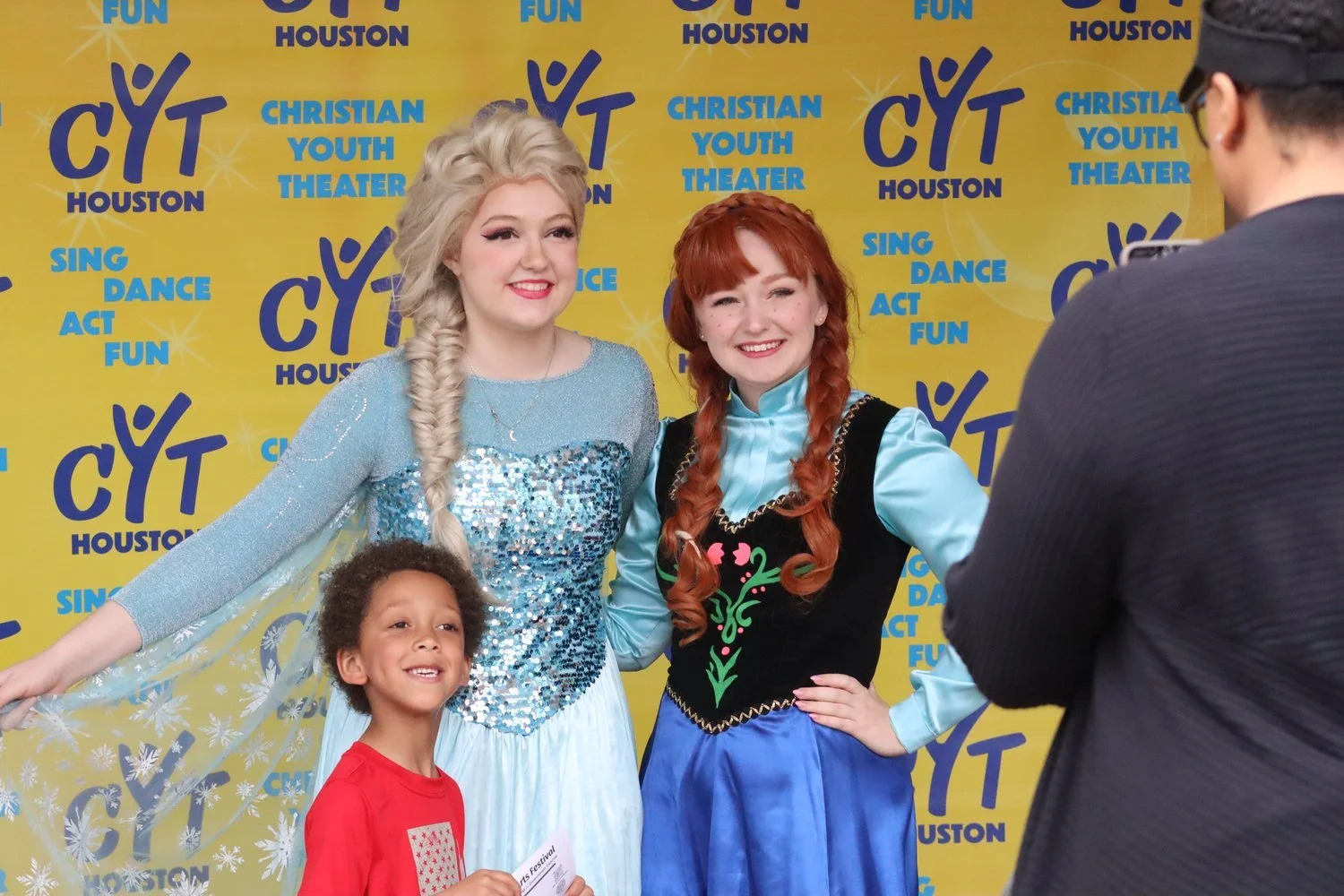 Two women and a young boy standing together at an event with a yellow backdrop that reads 'CYT Houston' and 'Christian Youth Theater' and lists activities like sing, dance, act, fun. The women are dressed as characters Elsa and Anna from Disney's Frozen, smiling for a photo being taken by a man.