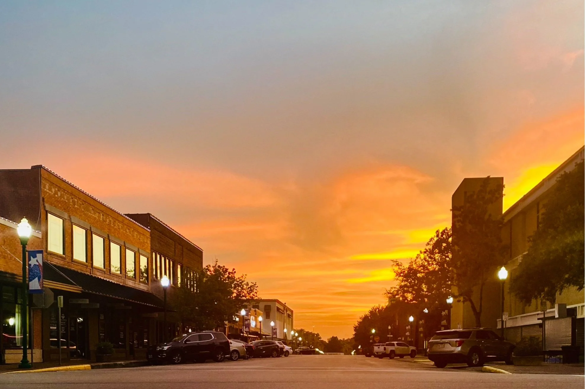 Empty street in a small town during a colorful sunset with buildings, trees, parked cars, and street lamps.