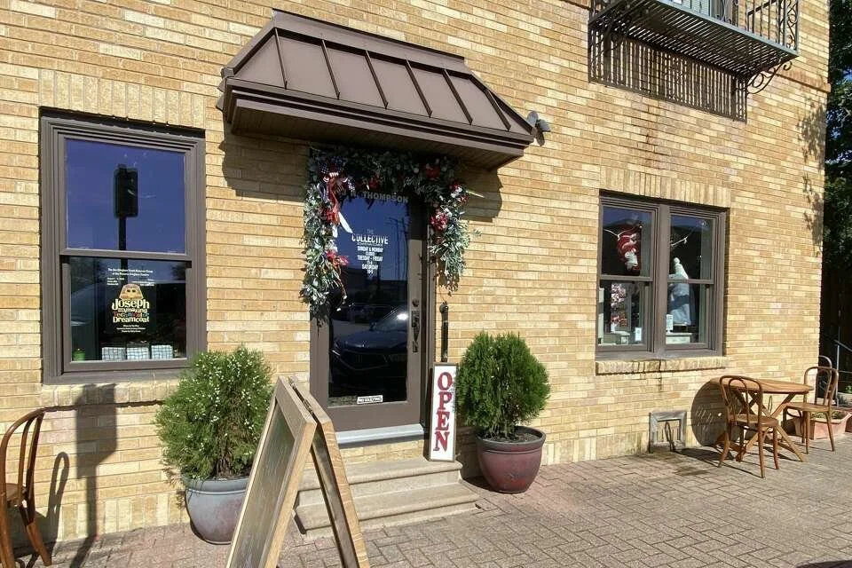 Exterior of a storefront with a glass door decorated with a holiday wreath and an 'OPEN' sign, two potted bushes on either side, and outdoor seating with three chairs and a table.