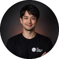 A young man with dark hair and a slight beard, smiling, wearing a black shirt with a logo on the chest, standing against a dark background.