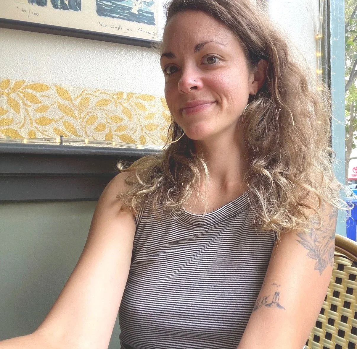 A young woman with curly brown hair and tattoos on her arms is sitting in a café near a window, smiling subtly at the camera.