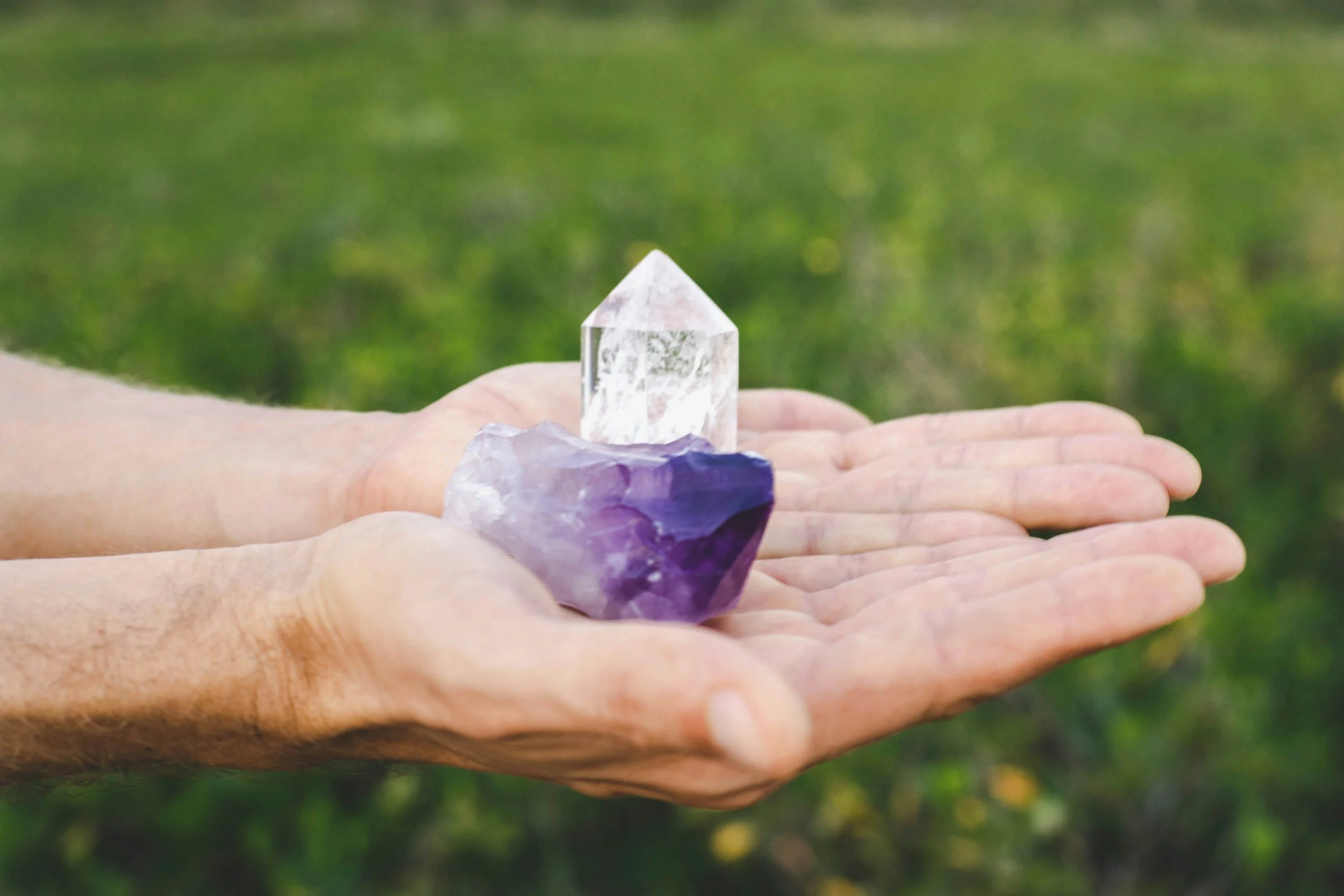 A person's hand holding a clear quartz crystal pointed like a tower, with a piece of amethyst crystal nearby, against a blurred green background.