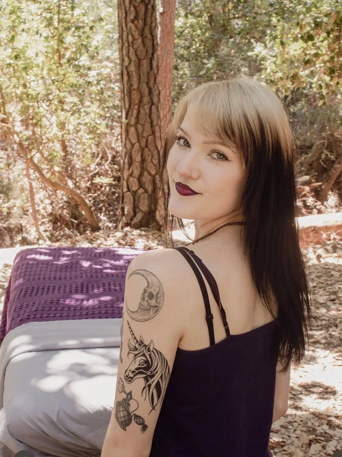 Young woman with tattoos on her arm, dark lipstick, and a septum piercing sitting outdoors among trees, smiling and looking over her shoulder.