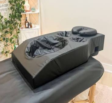 A black massage or medical treatment chair with a removable face cradle and cushioned arm supports, positioned in a treatment room with a small shelf holding bottles and a potted plant in the background.