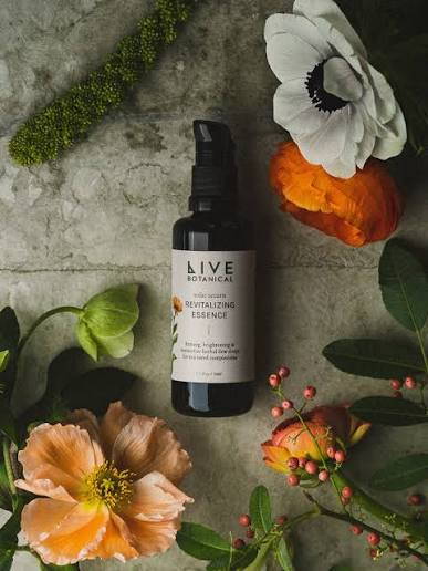 A black spray bottle labeled 'Live Botanical Revitalizing Essence' surrounded by various colorful flowers and green leaves, set on a concrete surface.