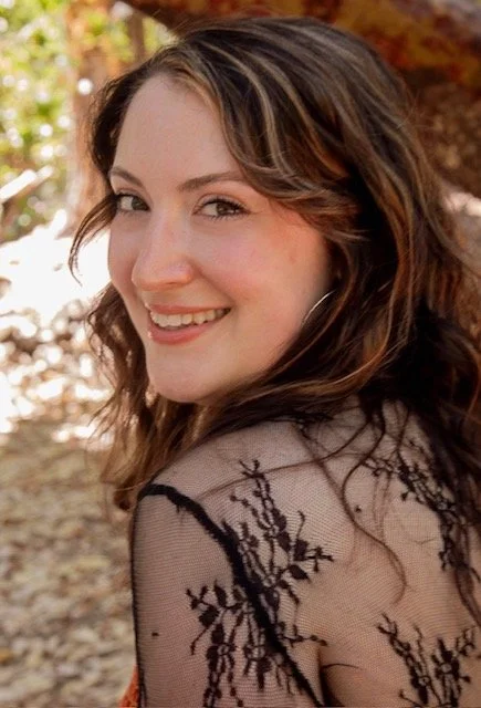 A young woman with wavy brown hair smiling outdoors with trees and autumn leaves in the background.