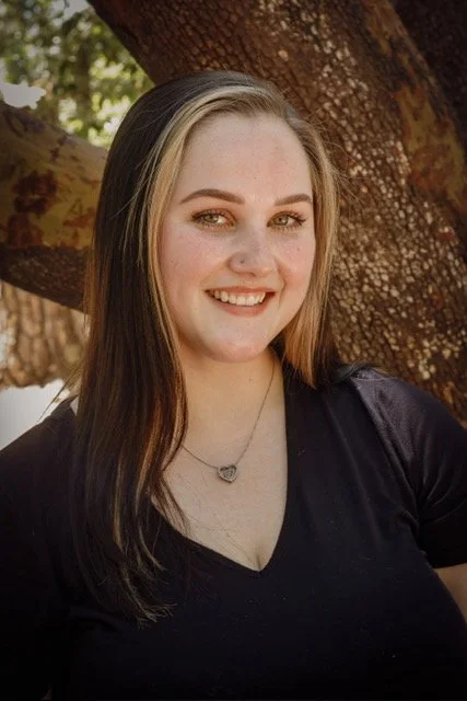 A young woman with long brown hair, wearing a black shirt and a silver necklace, smiling outdoors in front of a tree.