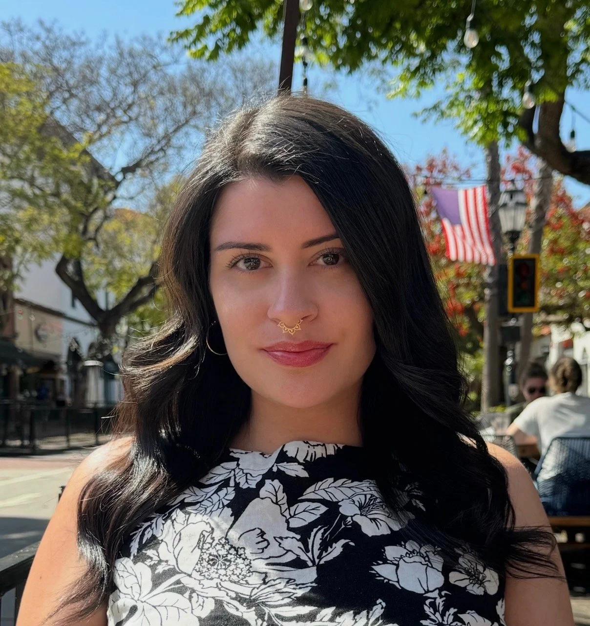 A woman with black hair, wearing a gold septum piercing and a black and white patterned sleeveless top, sitting outdoors during daytime with green trees, a traffic light, and an American flag in the background.