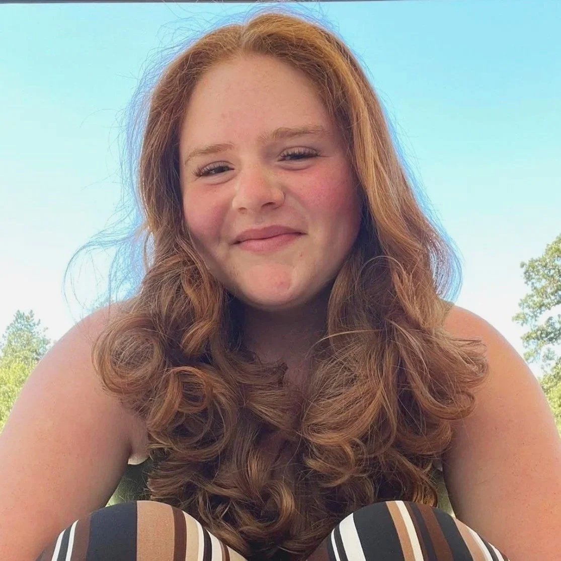 A young girl with long, curly red hair smiling outdoors with a blue sky and trees in the background who is a massage therapist specializing in deep tissue and recovery techniques.