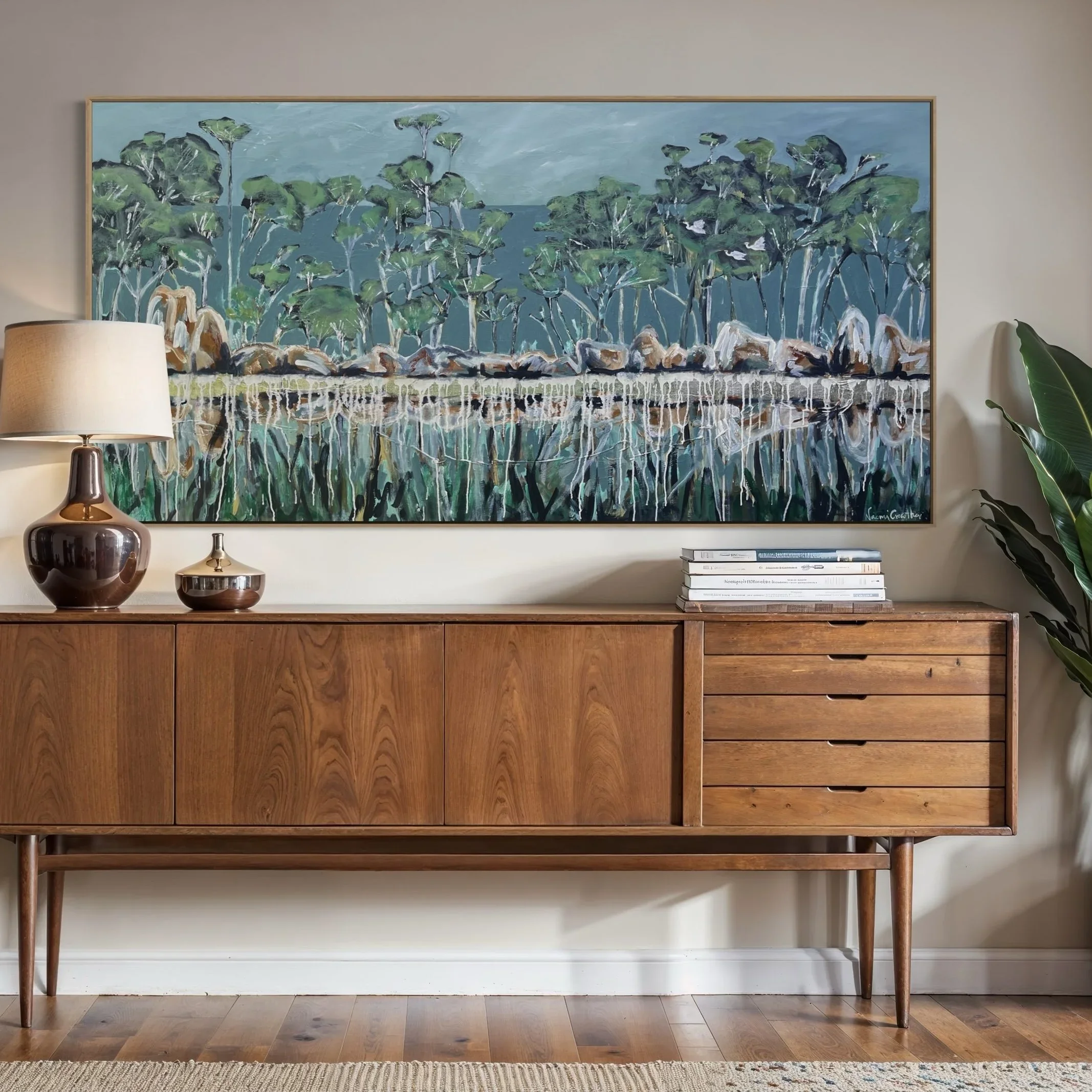 A wooden sideboard with three drawers and four legs, placed against a light-colored wall. On top of the sideboard, there are two decorative vases, one taller and one smaller, both with a shiny, reflective surface, and a small green plant in a pot on the right side. Above the sideboard, a large abstract painting of trees with green foliage and a blue background hangs on the wall.