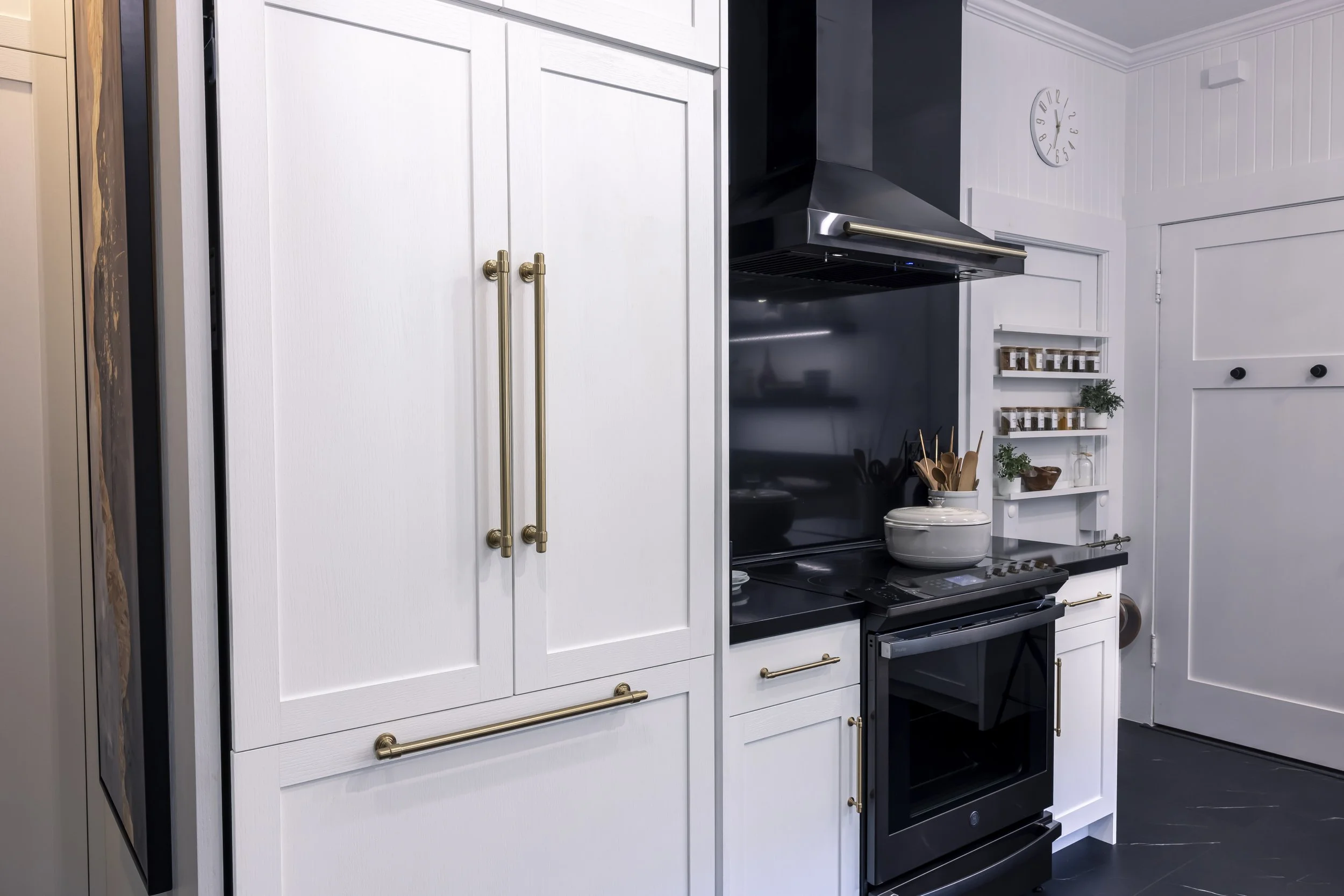 White kitchen cabinetry with integrated refrigerator, black range and backsplash, brass hardware, and custom spice storage.