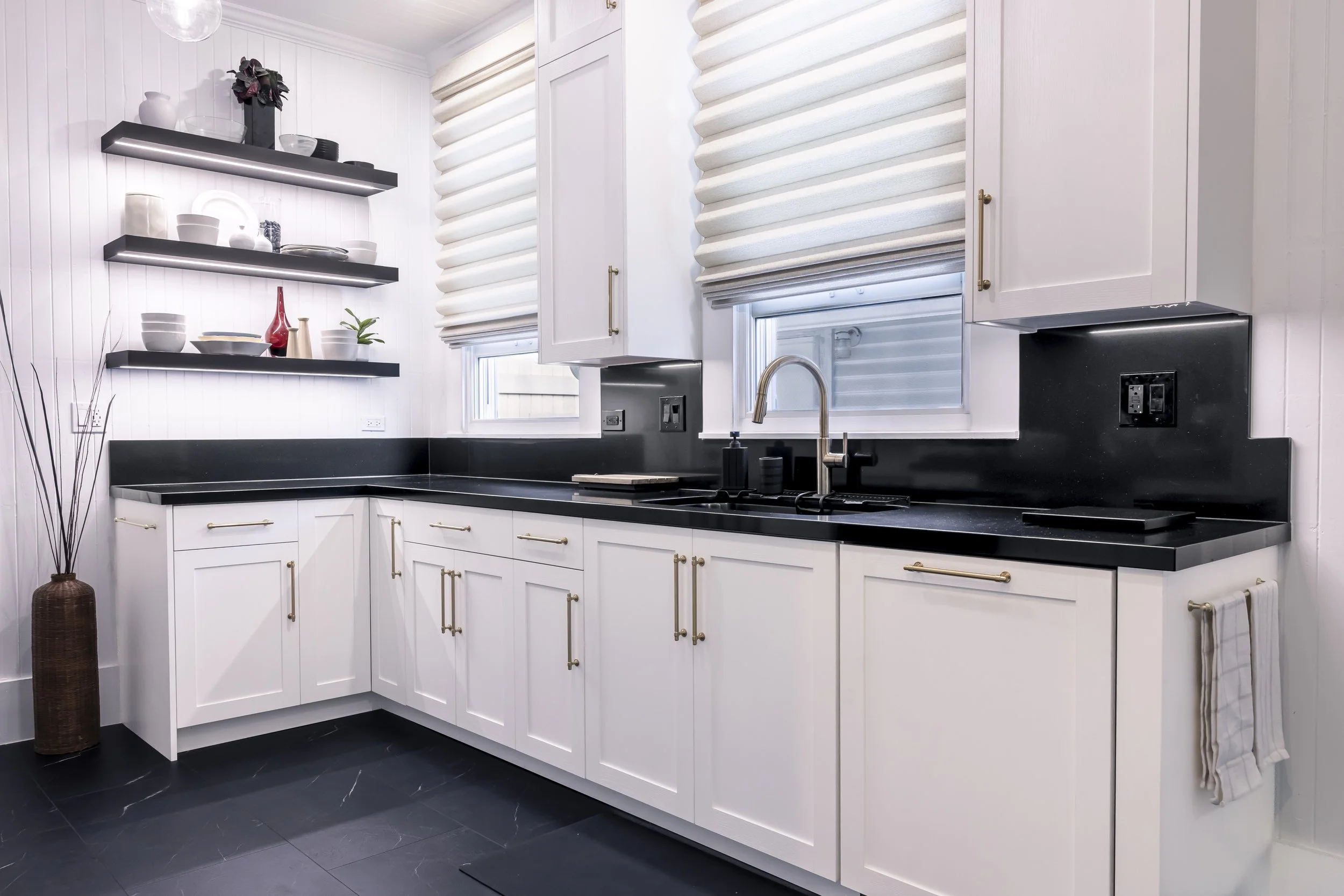 Bright Manoa kitchen with white cabinetry, black countertops, brass hardware, preserved windows, and illuminated floating shelves