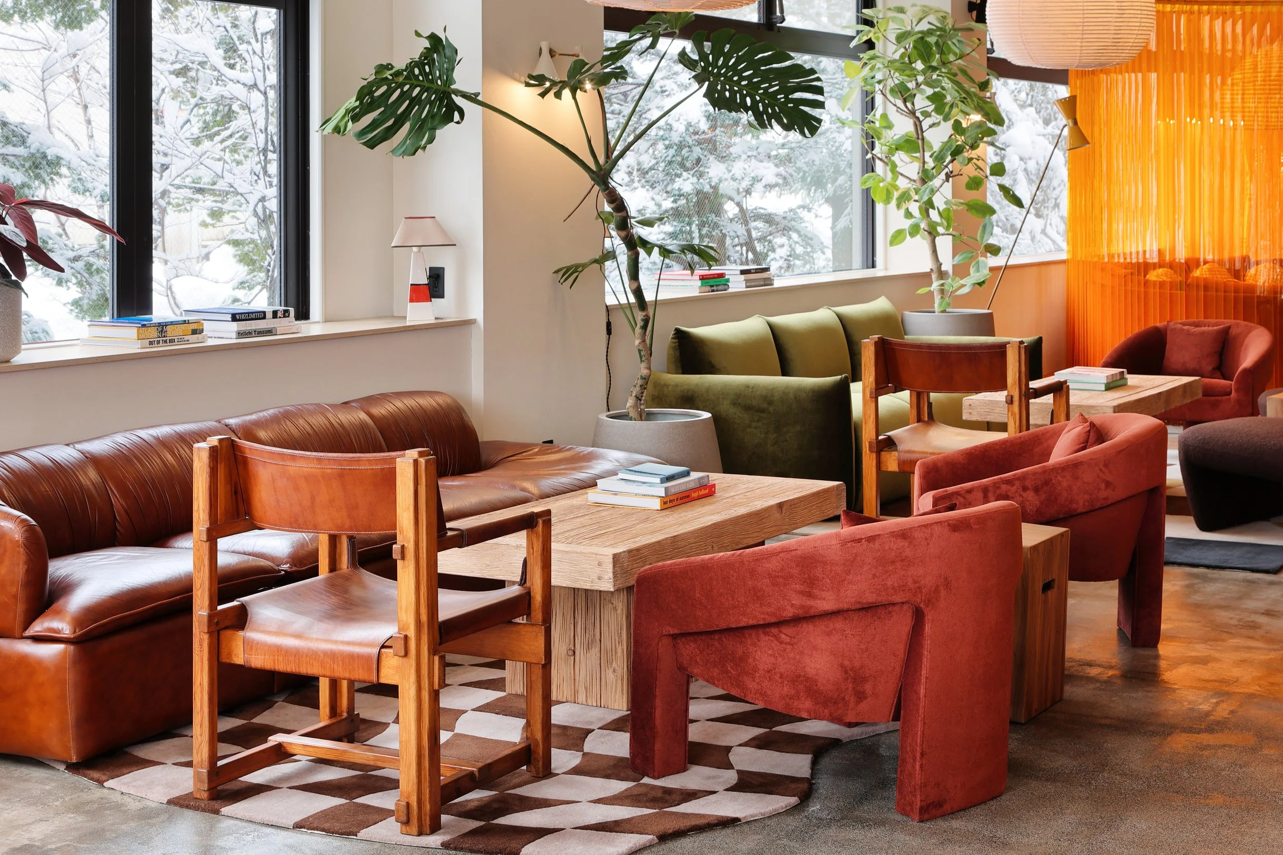 A cozy indoor lounge area with a leather couch, colorful velvet chairs, wooden tables, large indoor plants, and snowy landscape visible through large windows.