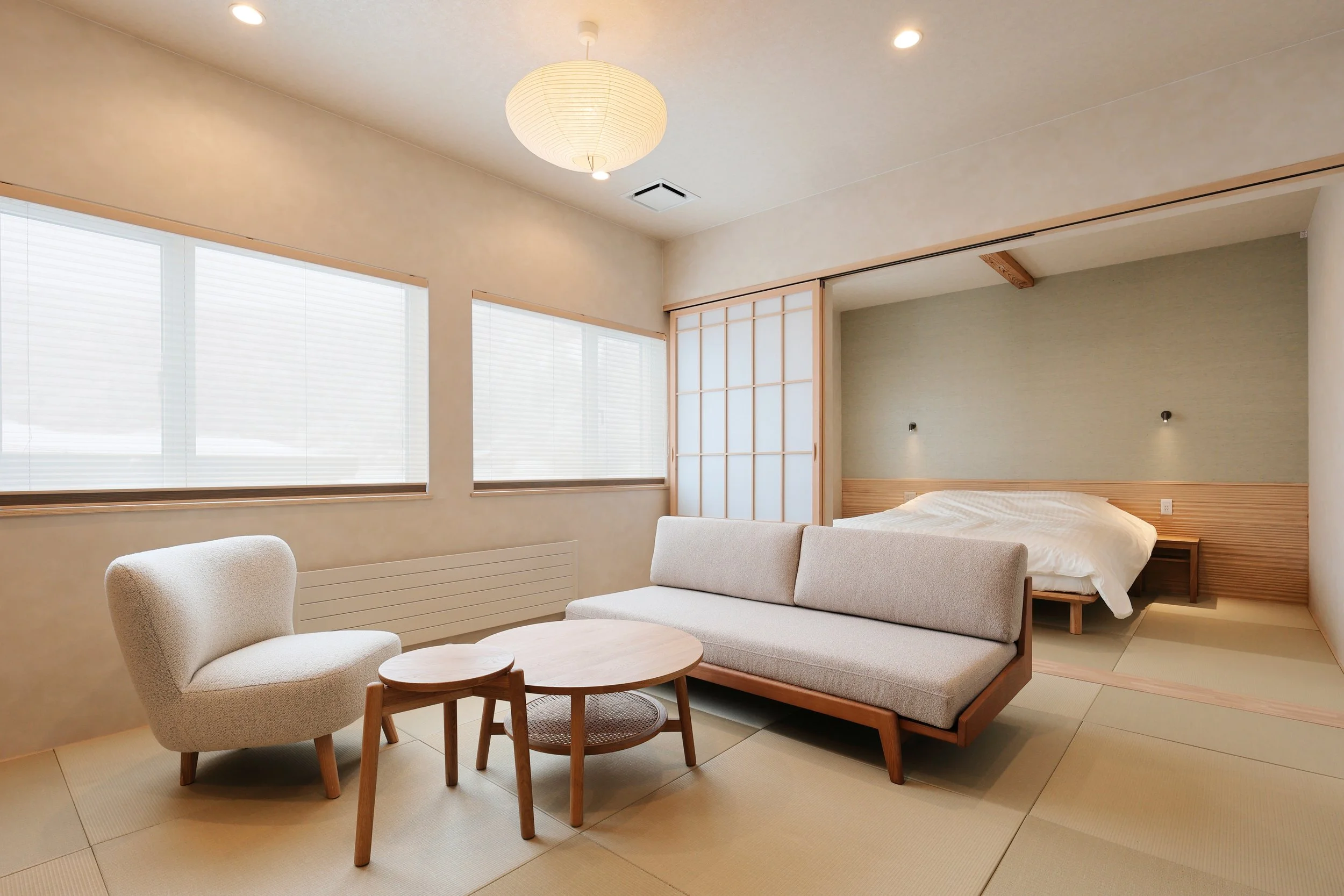 Minimalist Japanese-style room with tatami mats, a low bed, a sofa, a round coffee table, and large windows with blinds.