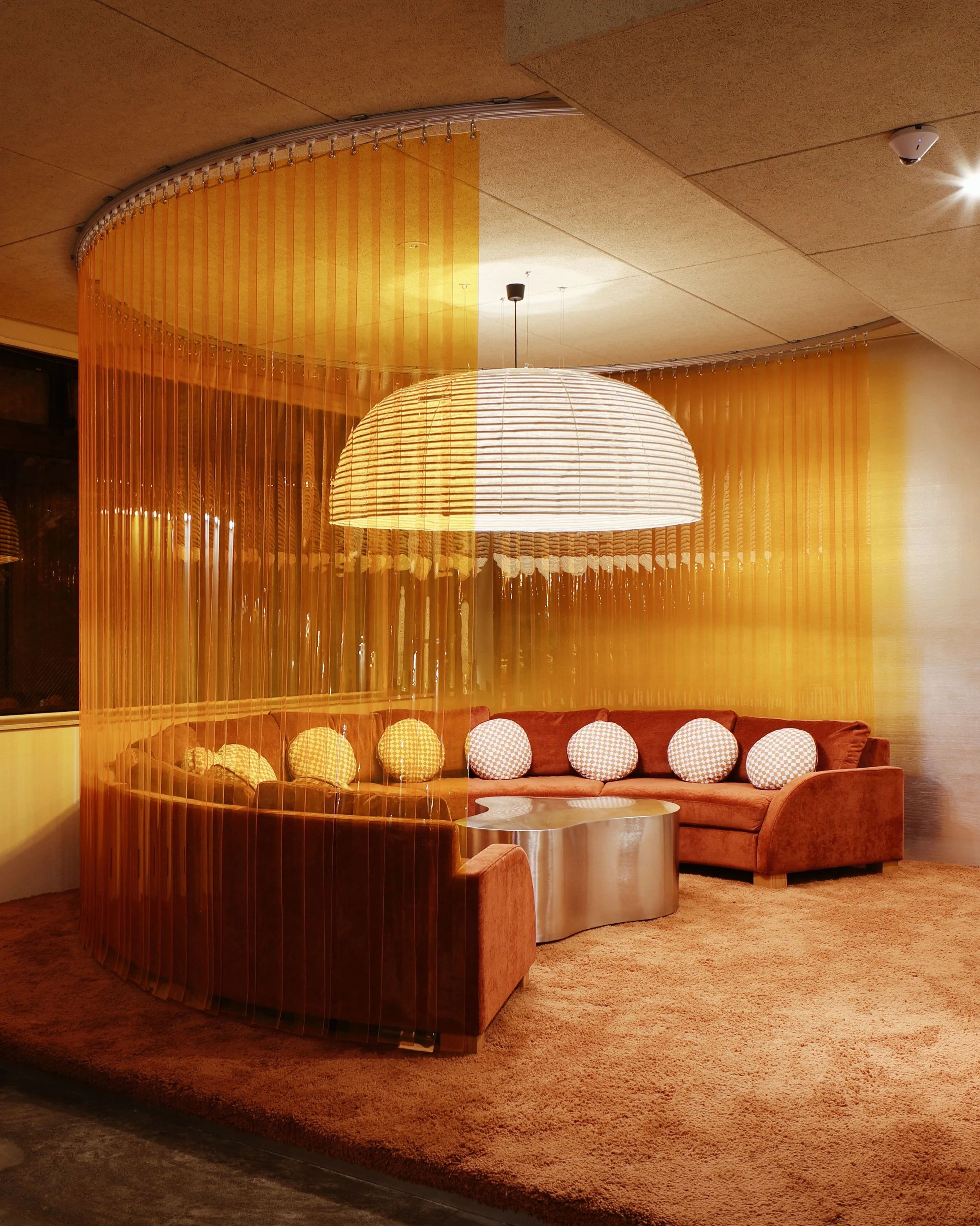 A cozy seating area with a curved, brown velvet sofa adorned with multiple round white and orange patterned pillows. A shiny metallic coffee table sits in front of the sofa. A large, hanging paper lantern-style light fixture is centered above the area. The space is enclosed by semi-transparent, orange vertical curtain panels, creating a semi-private nook. The floor is covered with a thick, textured brown carpet.