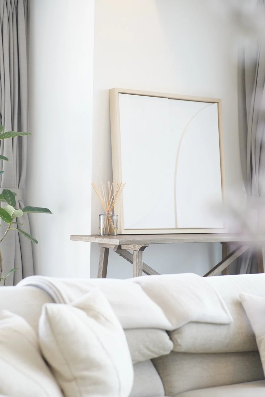 Decorative living room corner with a beige sofa, a wooden console table, a large framed minimalist art print, a small glass container with reed diffusers, and a green houseplant, with curtains on the window.
