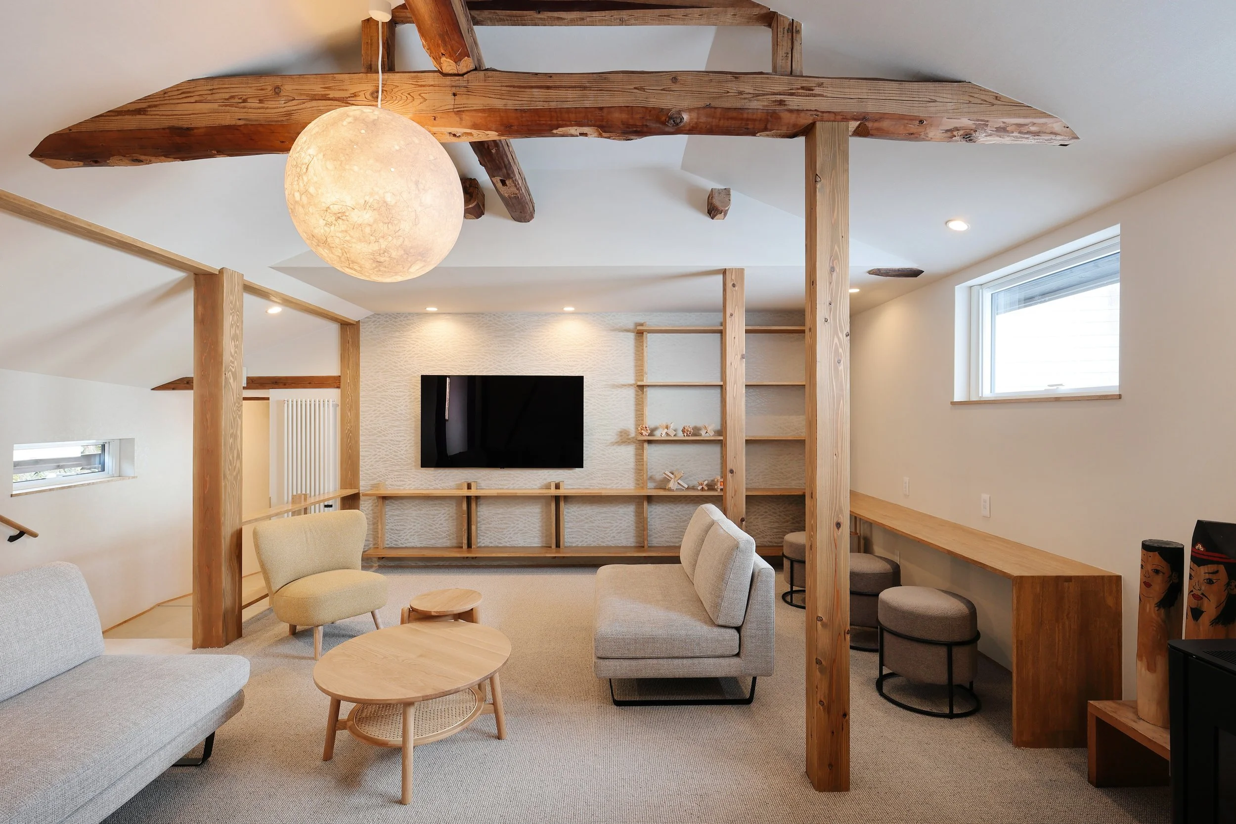 Modern living room with wooden beams, a large pendant light, a wall-mounted TV, beige sofas, a small round coffee table, a yellow armchair, a built-in wooden desk with stools, and a textured white accent wall. Small windows are on the side walls.