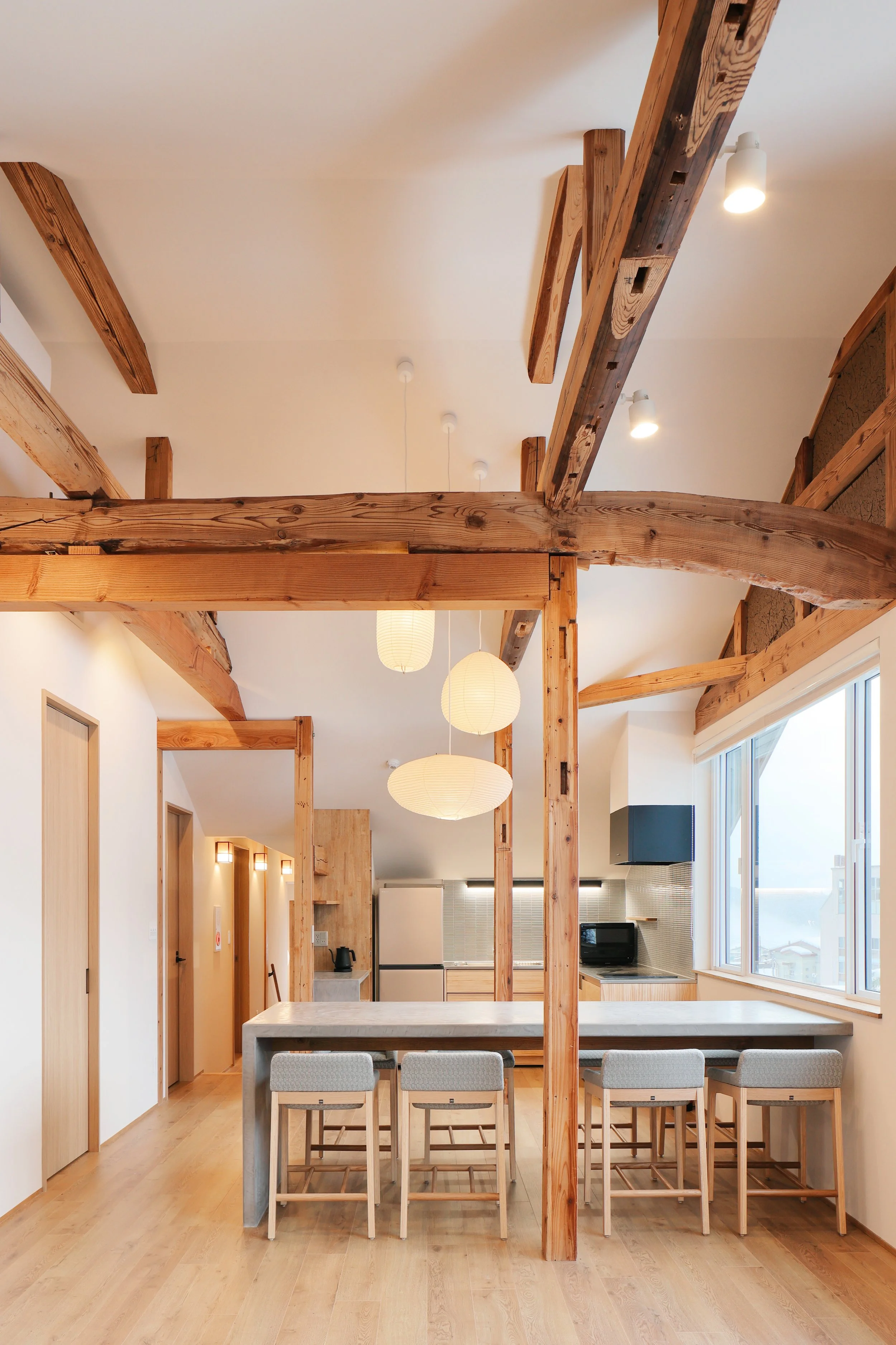 Modern kitchen and dining area with exposed wooden beams, pendant lights, a large window, and seating around a kitchen island.