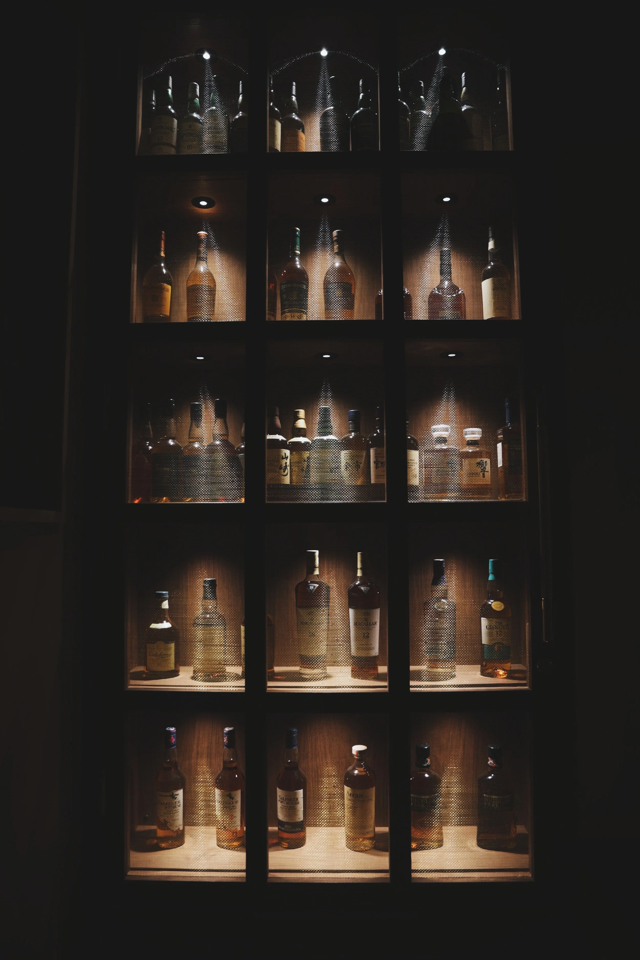 A display case with multiple shelves of liquor bottles, illuminated with small spotlights, against a dark background.