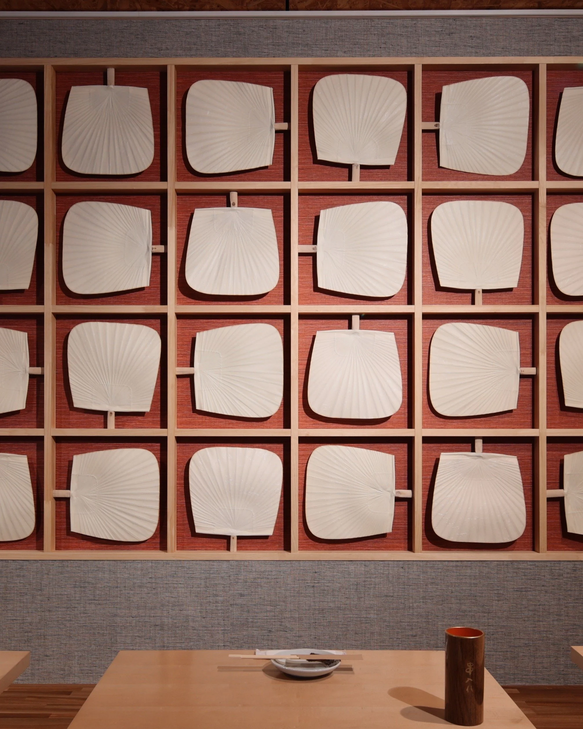 A wall decorated with a grid of 20 square wooden frames, each holding a white paper fan. In front of the wall, a table with a white bowl and a wooden cup.