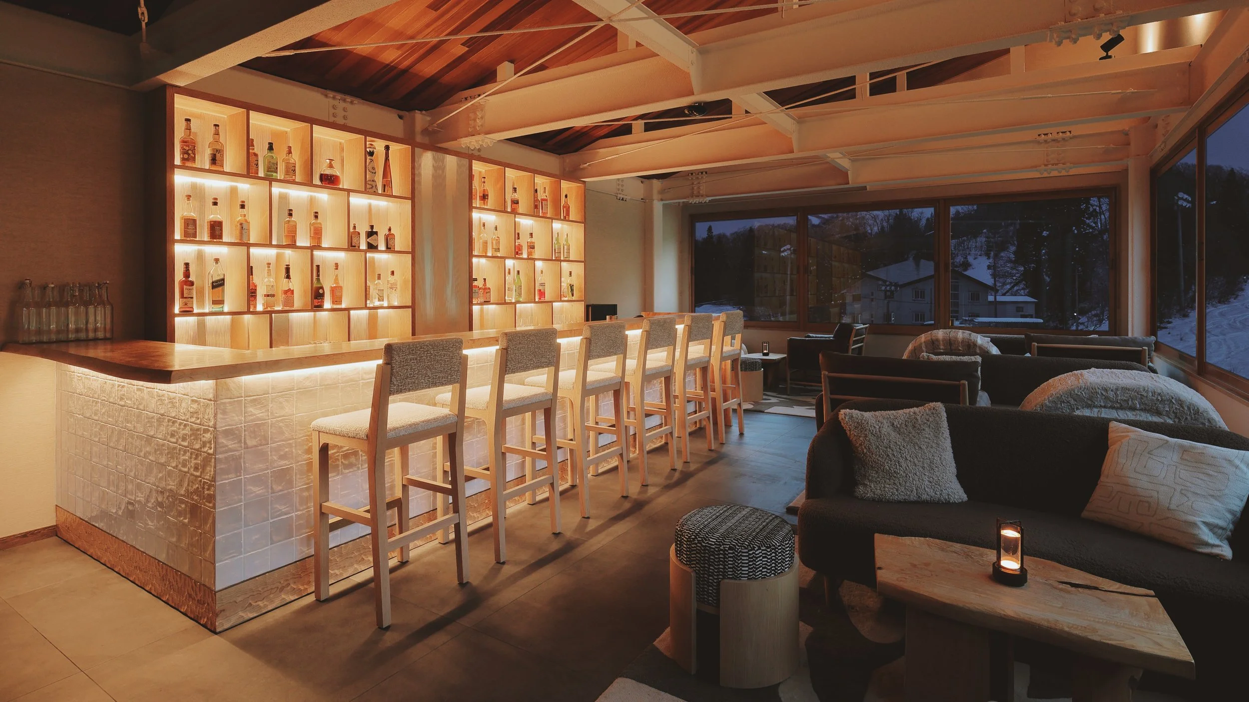 A cozy indoor lounge bar with warm lighting, a wooden bar counter with white upholstered bar stools, illuminated shelves with various bottles of liquor, and a seating area with dark sofas, pillows, and a small table with a lit candle. Large windows show a snowy outdoor scene at dusk.