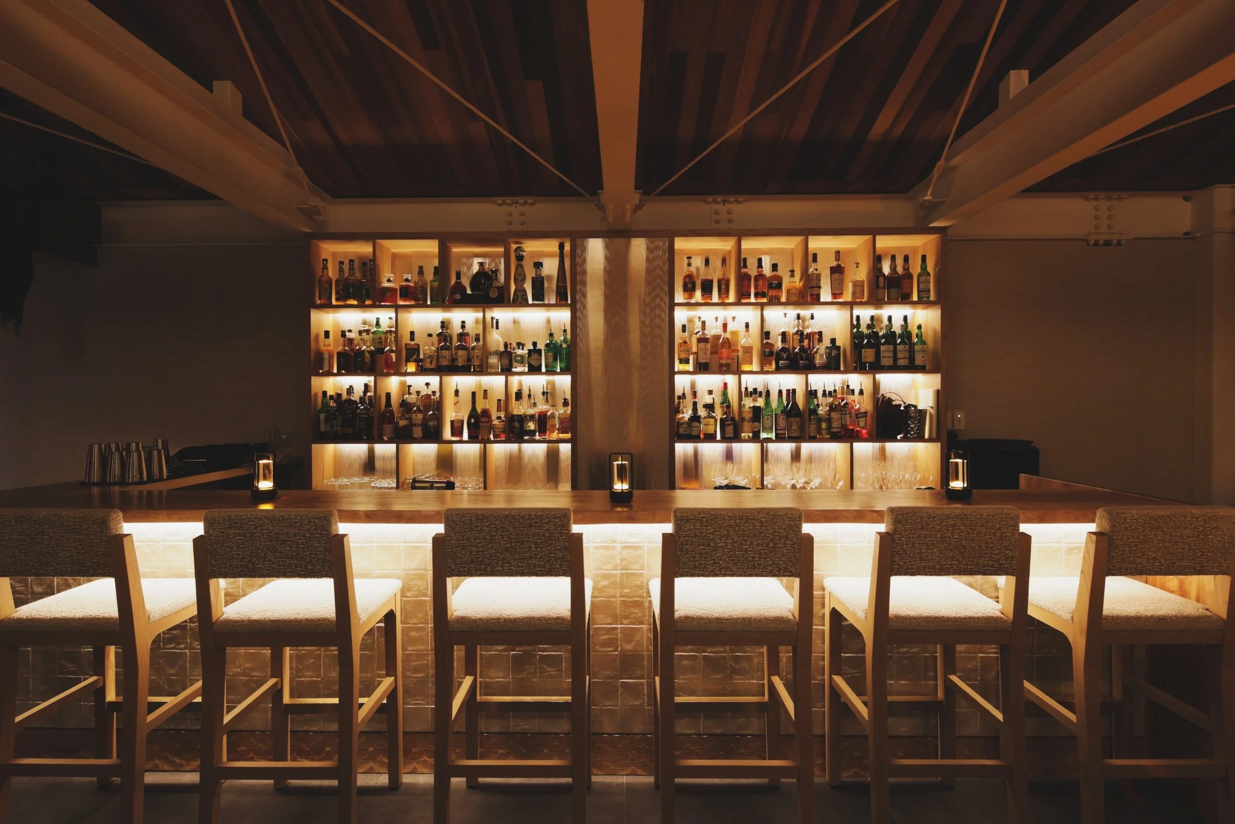 A cozy bar with a backlit shelf displaying various bottles of alcohol, a wooden counter with five chairs, and small lamps on the counter.