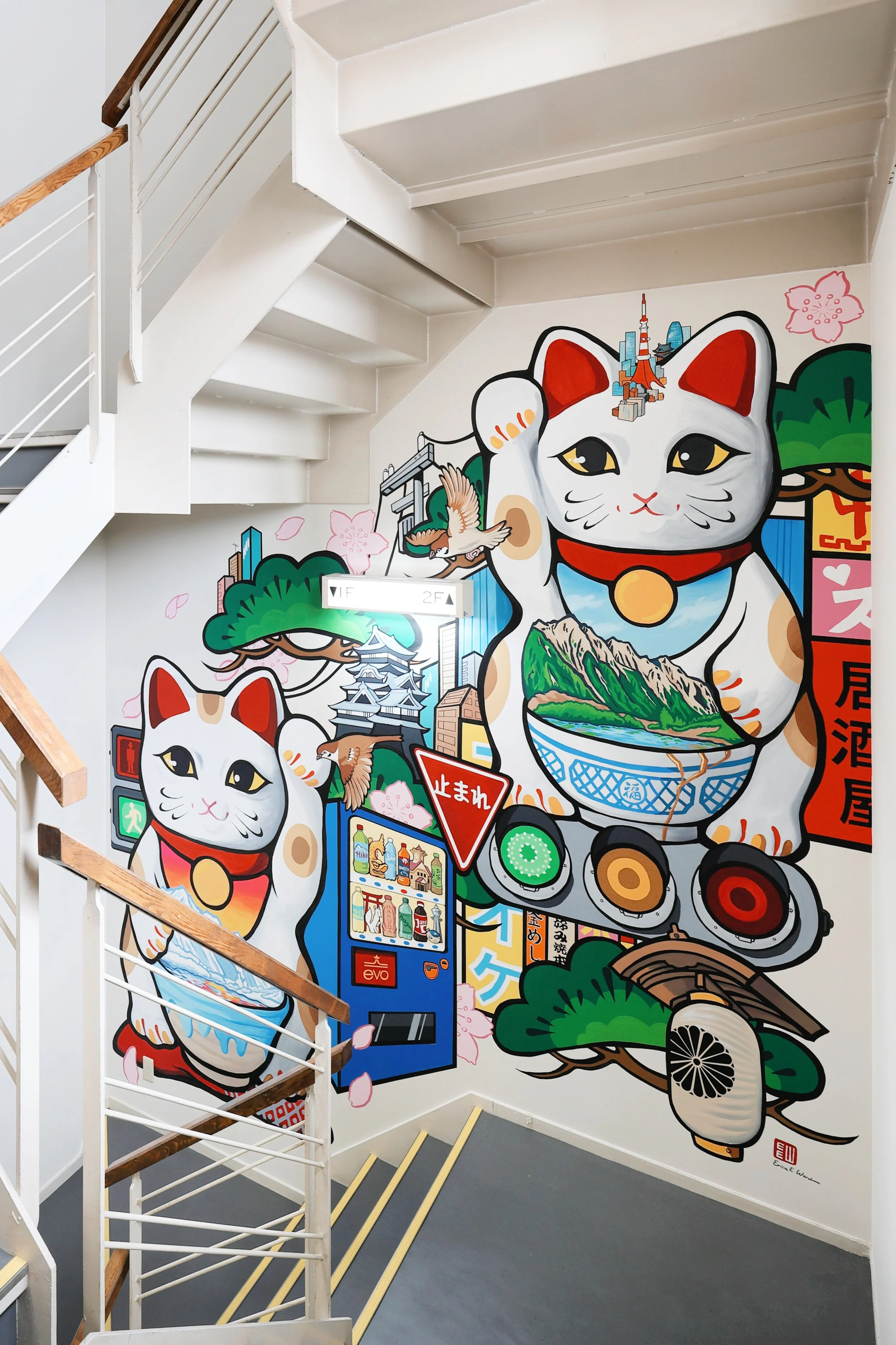Colorful mural of two Maneki-neko cats with a cityscape, Mount Fuji, cherry blossoms, and Japanese symbols, painted on a stairwell wall.