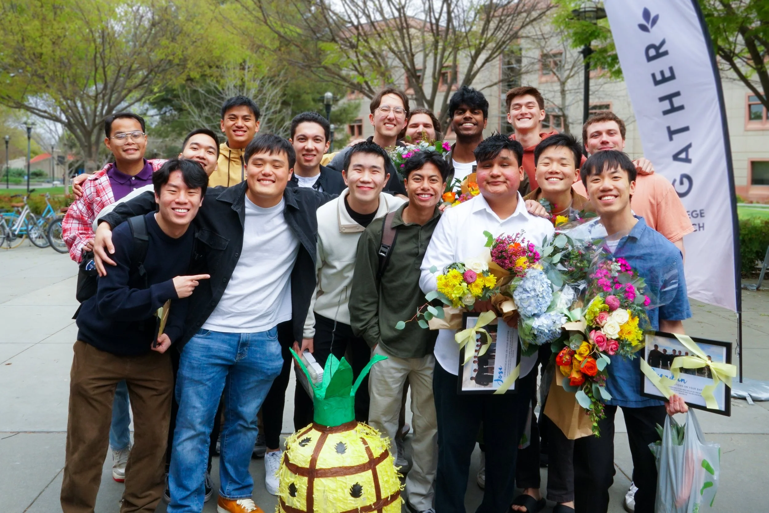 A group of UC Davis post grads smiling and posing outdoors on a college campus, holding bouquets of flowers, with trees and campus buildings in the background. This was after Baptism Sunday service. 
