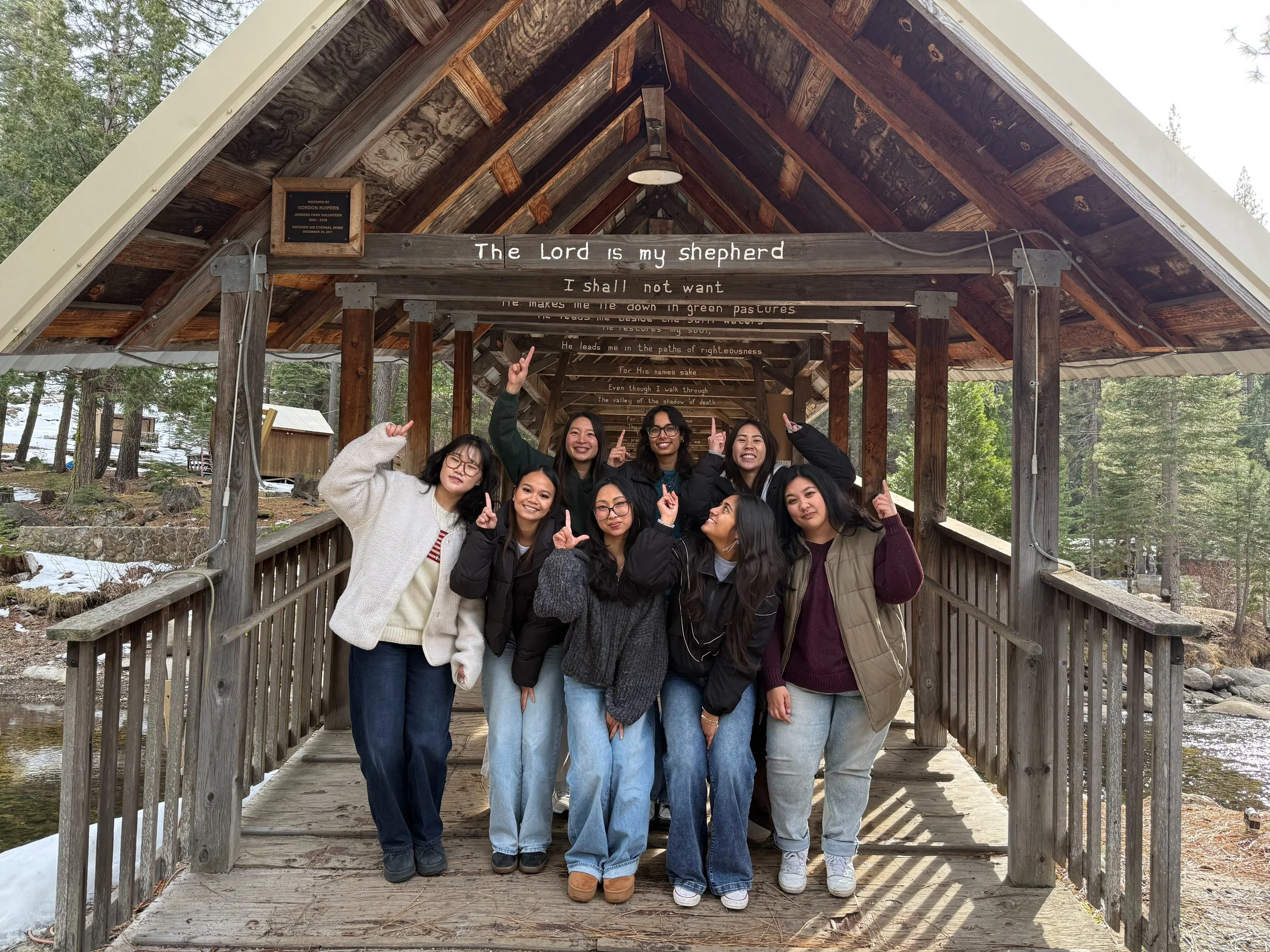 a group of UC Davis students standing together on a wooden bridge in a forested area, with snow on the ground, smiling and pointing upward after Christian Winter Retreat. 