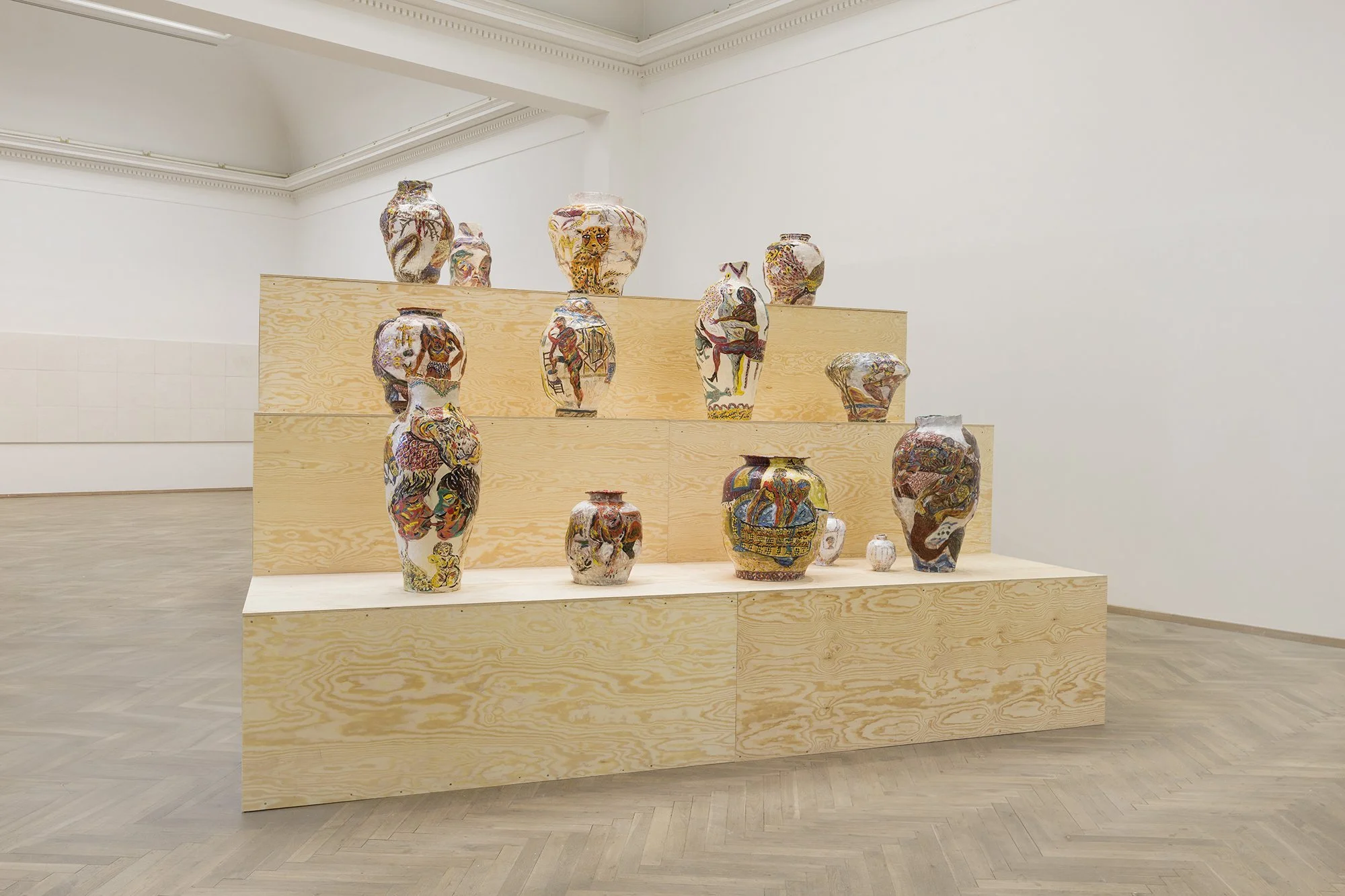 An art display featuring multiple colorful, painted vases on wooden tiered platforms in a gallery with white walls and wooden flooring.