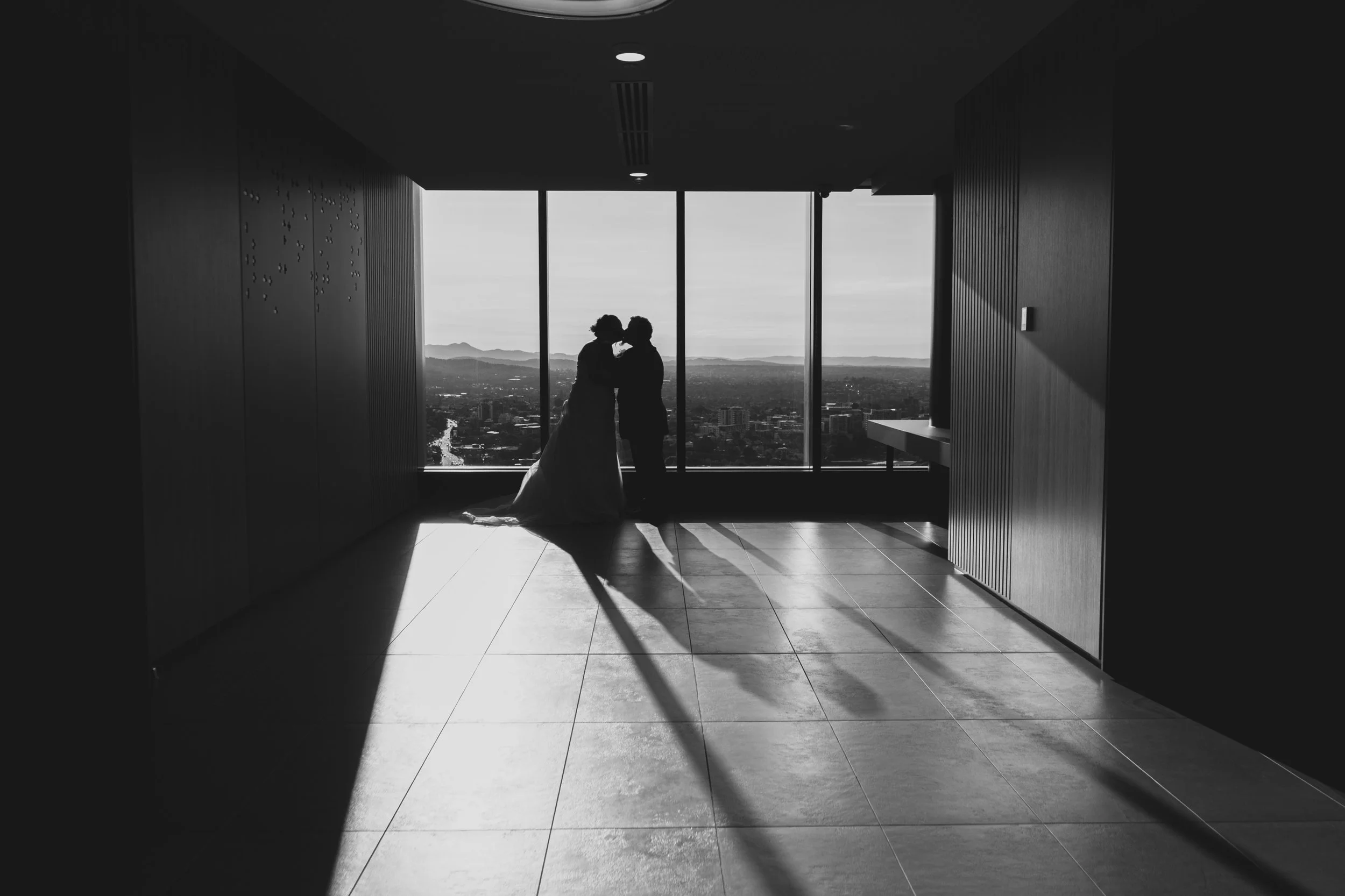 Silhouetted bride and groom standing close to each other near large windows, with cityscape and mountains in the background, in a modern indoor setting.