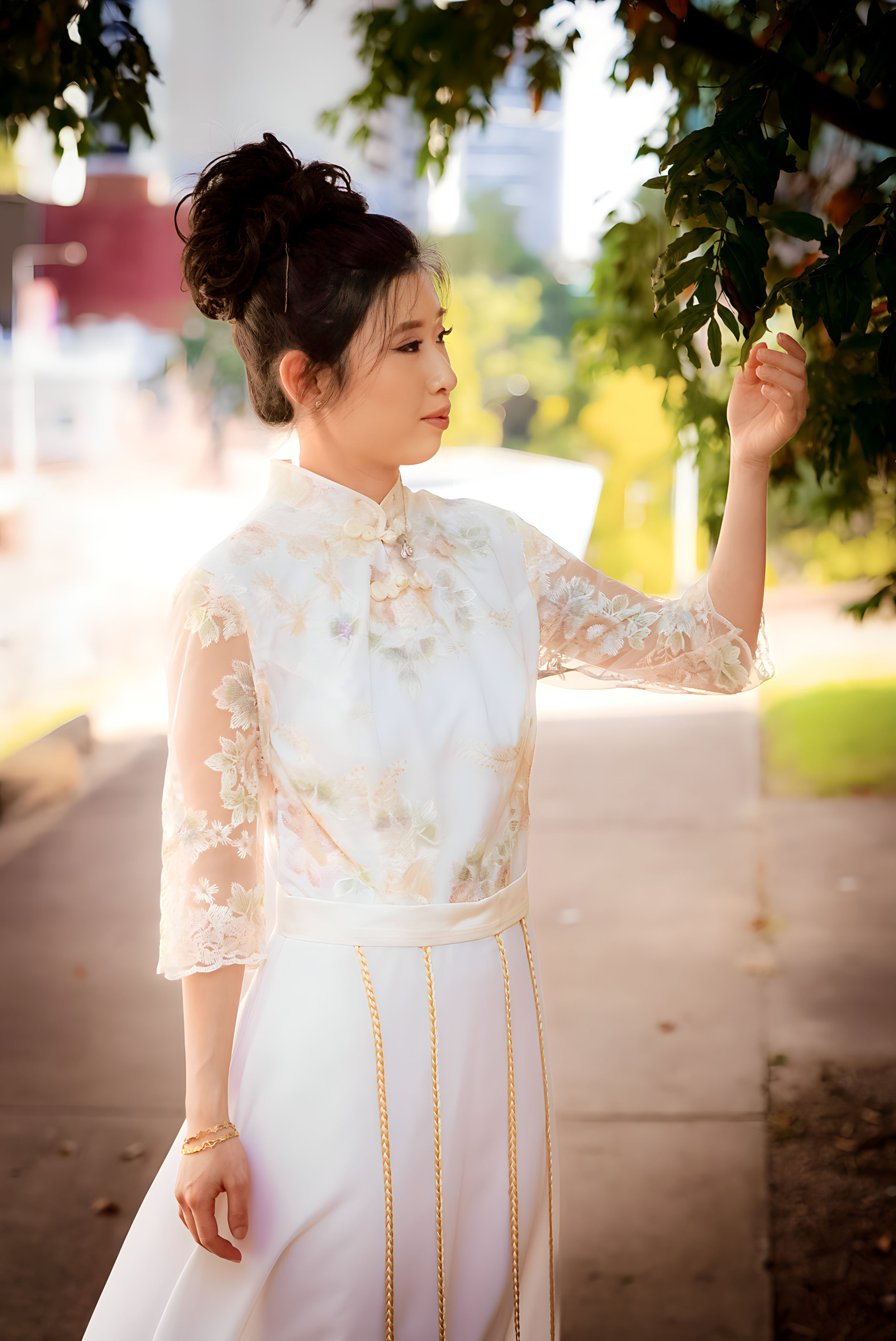 A woman wearing an elegant, embroidered white dress with lace sleeves, standing outdoors under tree branches, gazing at leaves.