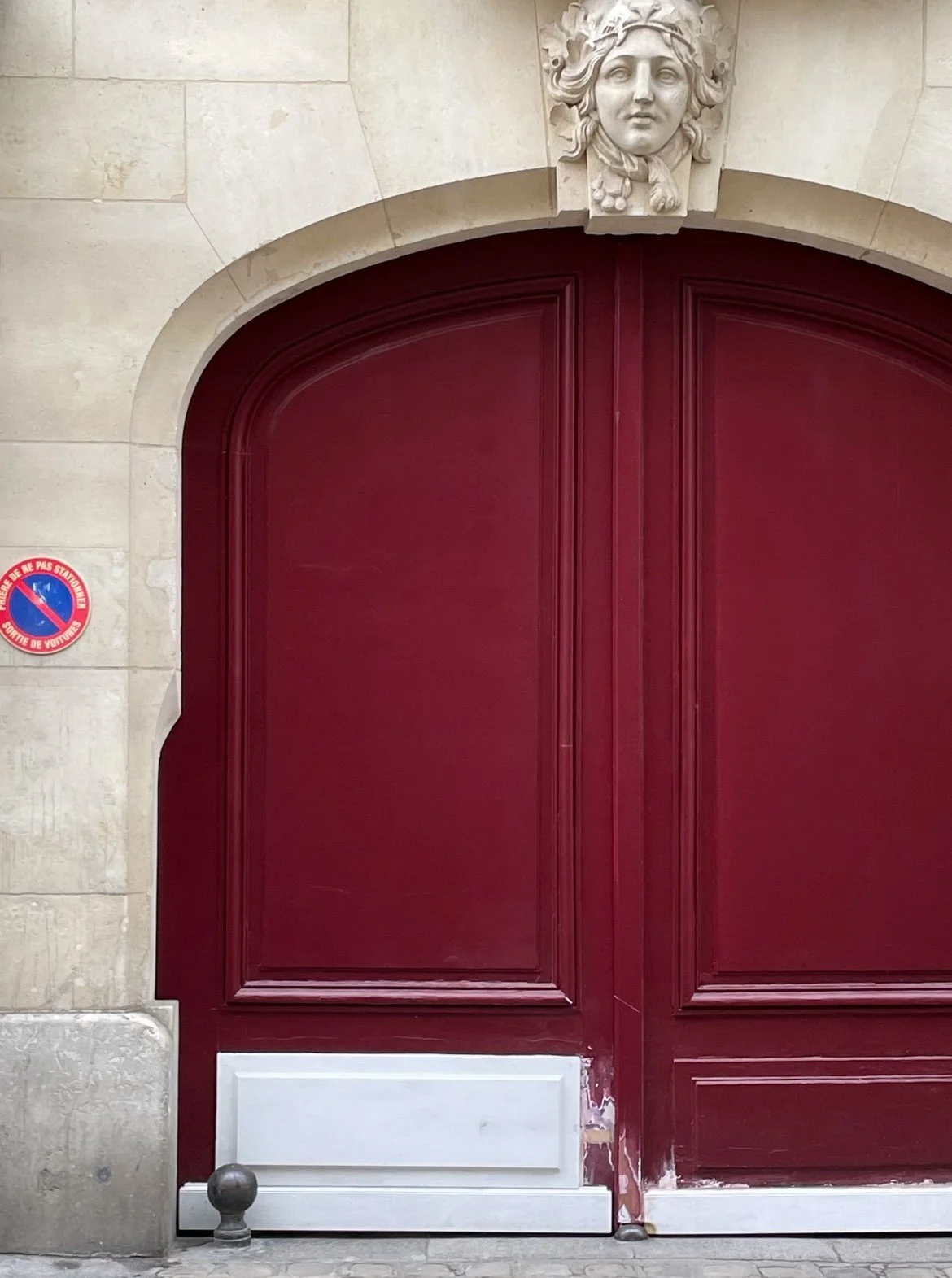 A large maroon double door with decorative molding. An Neoclassical style limestone sculpture of a face with a leafy crown is above the door. On the left side, there is a no parking sign in French on the stone wall.