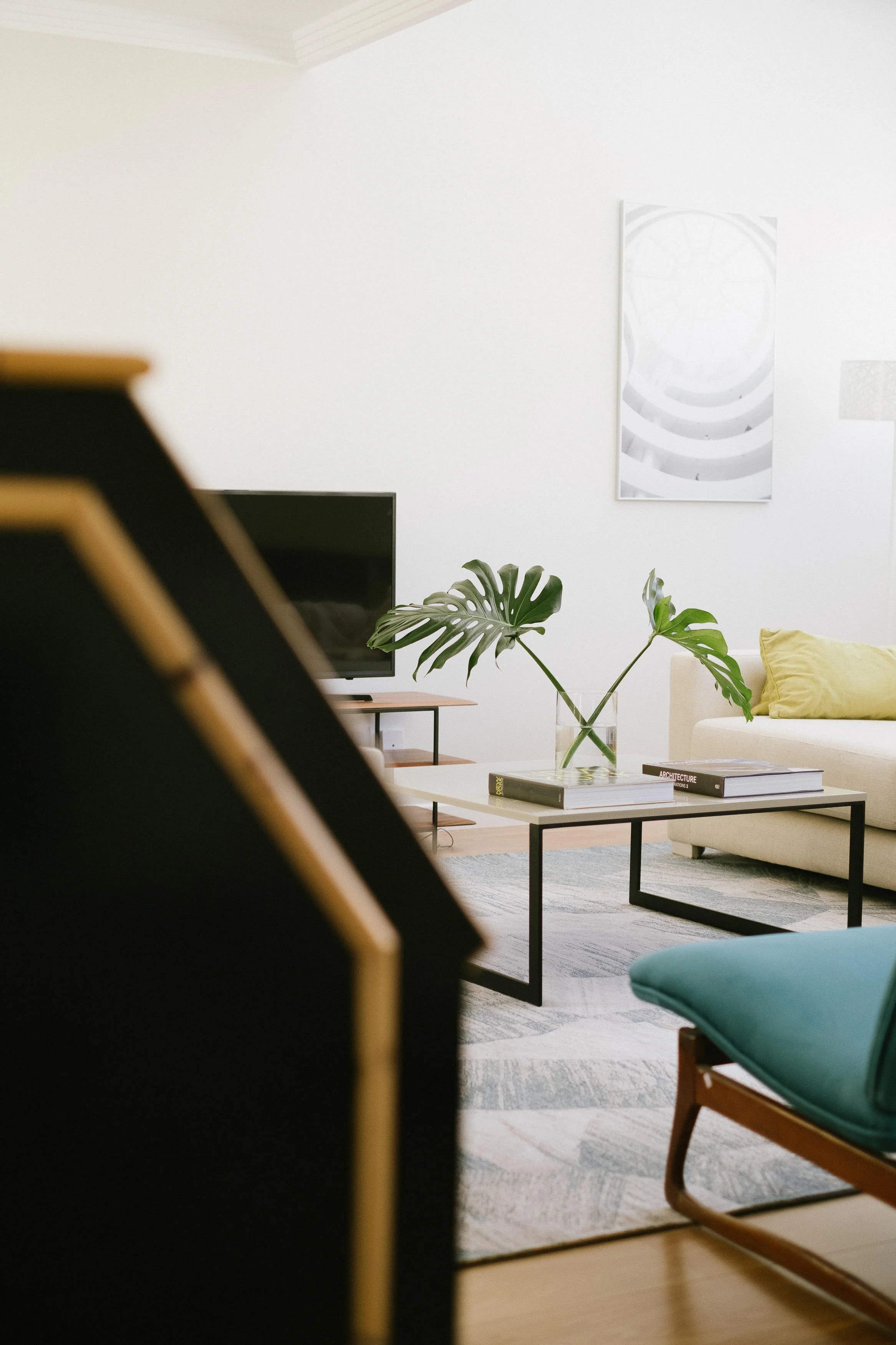 Living room with a black-framed television, a white coffee table with books, a green chair, a beige couch with yellow pillows, a large monstera plant in a glass vase, and wall art on a white wall.