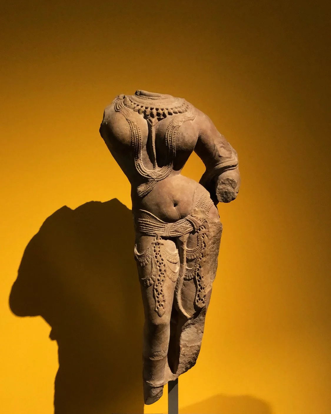 Ancient sandstone statue of a female figure decorated with jewelry, displayed against a yellow background.