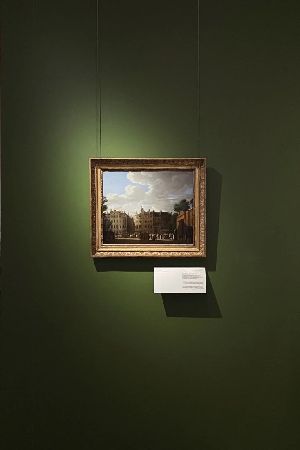 A framed painting of a cityscape hanging on a dark green gallery wall, with a white information plaque beneath it casting a shadow on the wall.