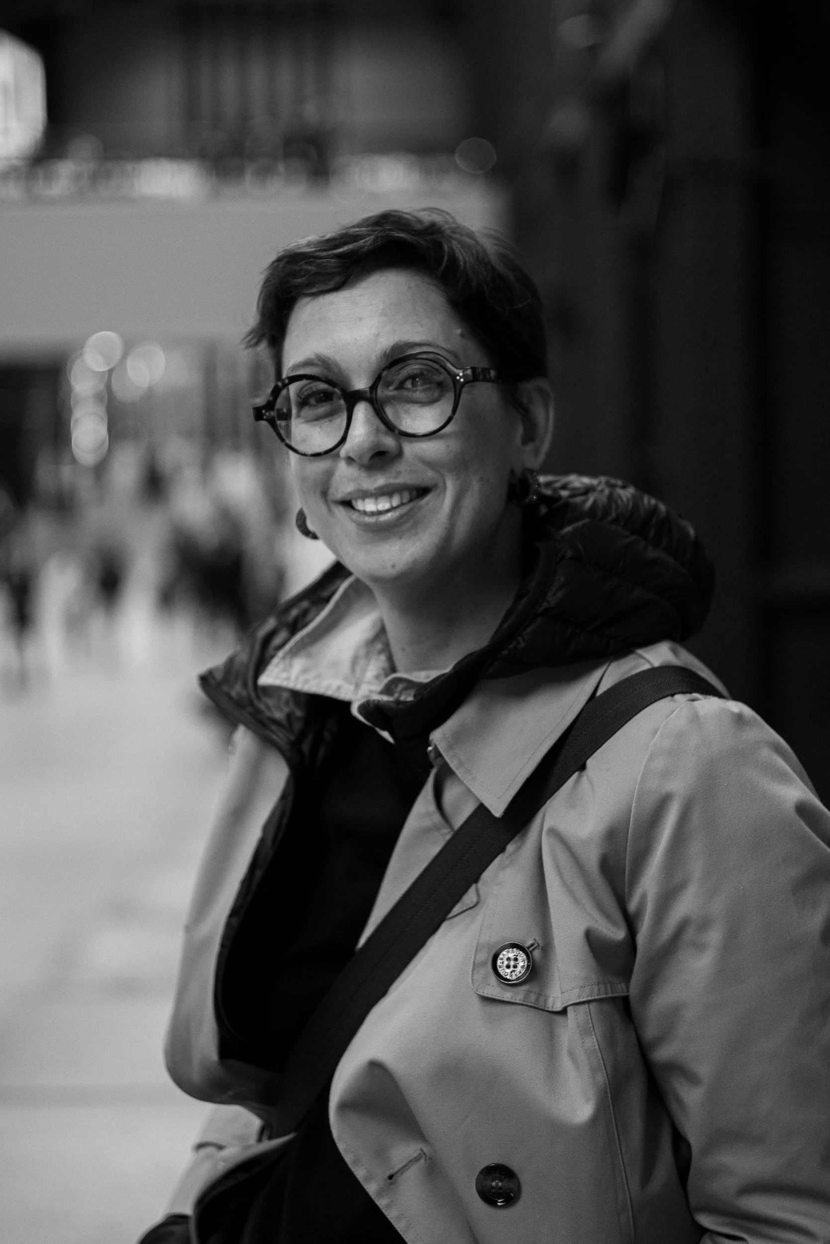 A woman wearing glasses and a light-colored jacket with a badge, smiling and looking at the camera in an indoor setting.