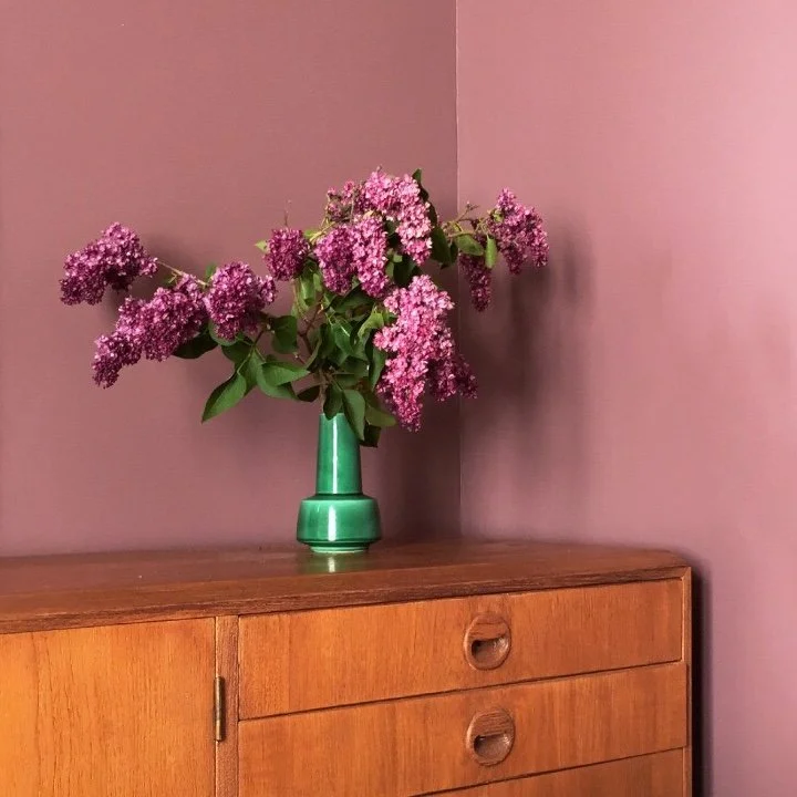 A bouquet of pink and purple lilacs in a green vase on a wooden cabinet against a pink wall.