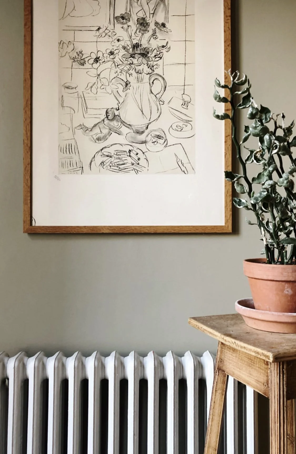 A framed sketch of a still life scene with flowers, a pitcher, a plate, and various objects on a table, hanging on a wall next to a potted plant on a wooden table and a white radiator below.